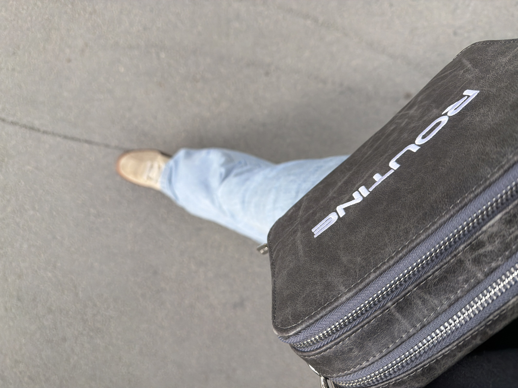 The image presents a high-angle, first-person perspective looking down at a person's legs, feet, and a bag. The setting is an outdoor, paved surface, likely concrete or asphalt, in what appears to be an urban or suburban environment, specified as Yekaterinburg, Russia.
The subject is a person, whose lower body is visible. They are wearing light blue jeans and a pair of light-colored shoes, possibly sneakers or canvas shoes, on their feet. A dark grey, rectangular bag with a textured finish is prominently displayed in the foreground, held or worn by the person. The bag features two visible horizontal zippers with metallic teeth and fabric pull tabs, suggesting multiple compartments.
The most notable detail is the white text emblazoned on the bag, clearly spelling out "ROUTINE" in capital letters. The lighting is bright and even, indicating daytime, although there are no strong shadows to suggest direct sunlight or a specific time of day. The activity captured is likely walking or standing, seen from the perspective of someone looking down at their own attire and belongings. No specific emotions are discernible. The scene conveys an everyday, casual moment.