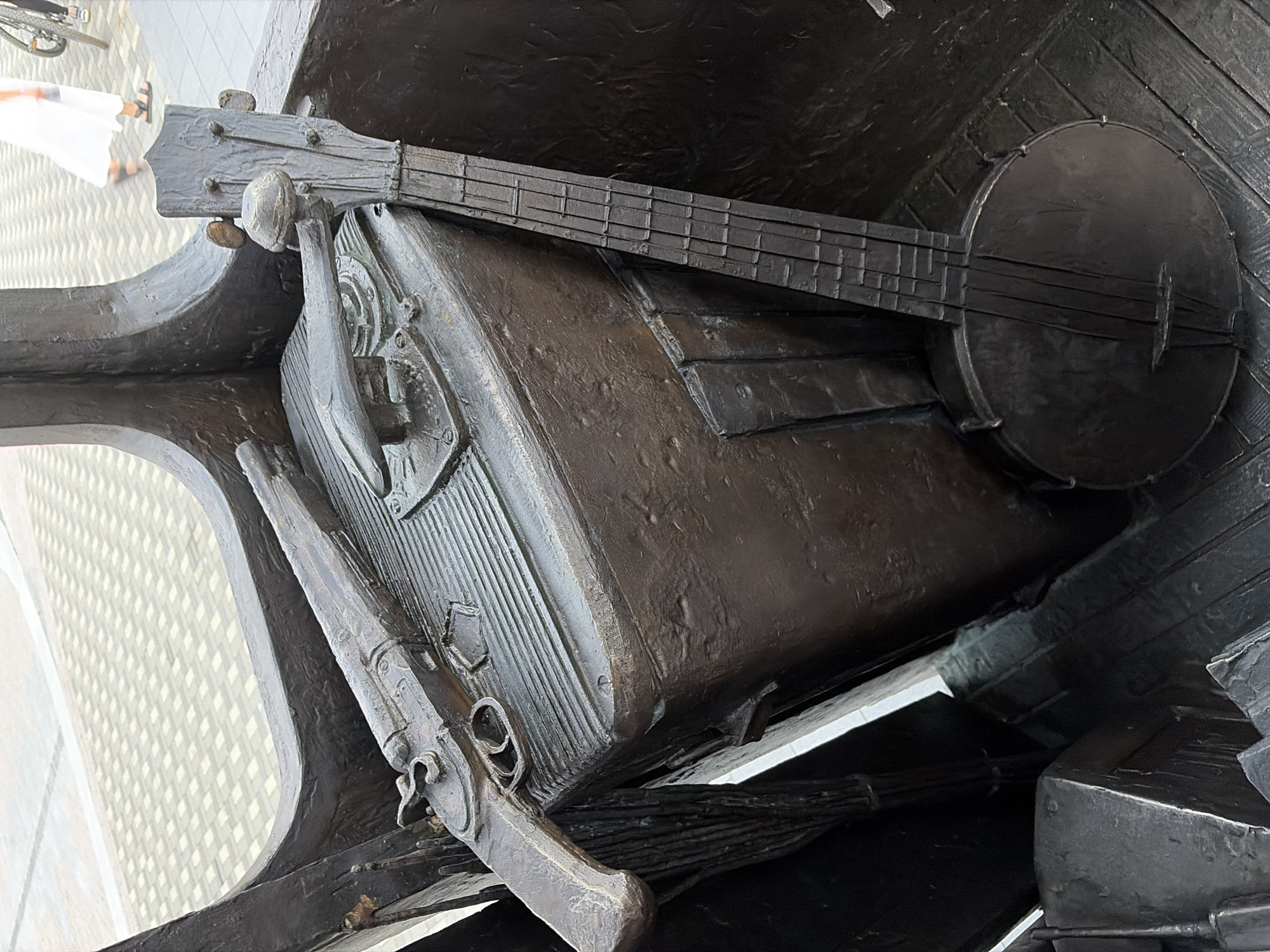 This is a close-up, eye-level, medium shot of a bronze sculpture depicting a vintage rifle and a banjo. The rifle, an antique flintlock style, rests horizontally across the top of a dark, rectangular object, possibly a trunk or a large case. The banjo is positioned vertically to the right of the rifle and case, with its circular body resting on a wooden plank-like surface. The sculpture appears to be part of an outdoor installation, as suggested by the tiled pavement visible in the blurred background and the legs of a person walking in the distance, indicating a public space. The setting is likely Yekaterinburg, Russia, where such artistic installations are common in urban areas.
