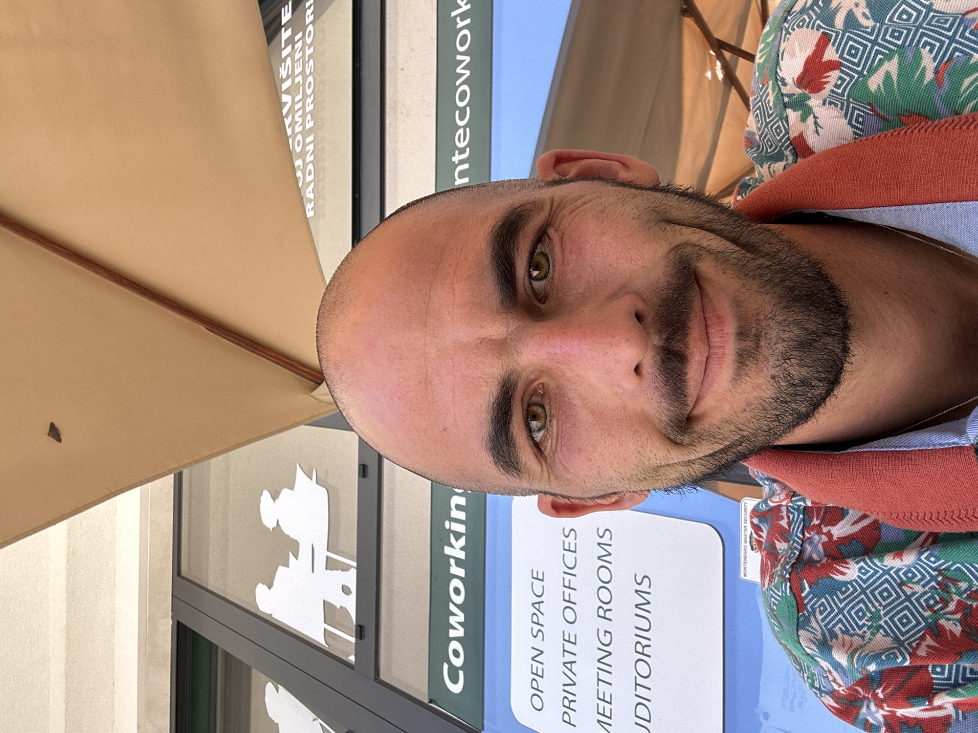A man is featured prominently in a close-up, selfie-style portrait, slightly smiling and looking directly at the camera. He has a bald head, a short dark beard, and light brown or hazel eyes. He is wearing a multi-colored patterned shirt with dominant blue, red, green, and white elements, and an orange collar or inner lining is visible around his neck.
The background reveals an outdoor or semi-outdoor setting, possibly a patio or a covered area, indicated by a large, light brown umbrella spanning the upper left corner. Behind the man, a modern building with large windows is visible. On the building, signs advertise a coworking space. Visible English text includes "Coworking", "OPEN SPACE", "PRIVATE OFFICES", "MEETING ROOMS", and "AUDITORIUMS". Another sign mentions "MONTENEGRO MASTER SECURITY". Text in a Slavic language, likely Montenegrin, Croatian, or Serbian, reads "OMILJENI RADNI PROSTORI" (meaning "Favorite Working Spaces"), with a partially visible word above it, possibly "KVIŠITE". Stylized white silhouettes of people sitting at desks are adhered to the glass windows.
The scene is brightly lit, suggesting it is daytime with clear weather. The image appears to be taken in Budva, Montenegro, consistent with the "MONTENEGRO" text. The overall impression is pleasant and modern, showcasing the man in front of a contemporary workspace.