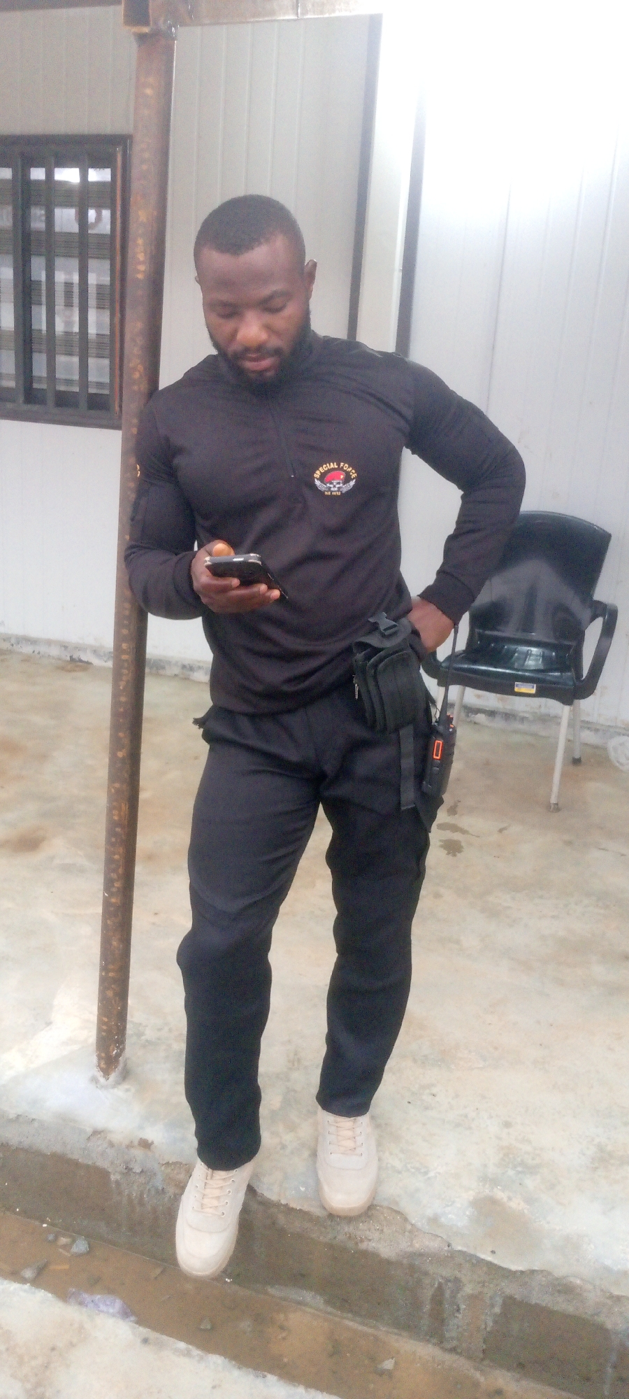 A full-body, vertical shot captures a dark-skinned man standing in what appears to be an outdoor setting, possibly the city of Indo, Nigeria. He is leaning against a rusty metal pole. The man is wearing a black, long-sleeved, quarter-zip shirt with a patch on the left chest that reads "SPECIAL FORCE." He also wears black pants with a utility pouch attached to his left hip. The pouch has a radio clipped to it. He holds a smartphone in his right hand, looking down at it. His feet are clad in light-colored sneakers. In the background, a black plastic chair is visible, and to the left, a window with horizontal slats is set into a light-colored wall. The ground is a mix of concrete and a dirt/gravel trench filled with murky water. The lighting suggests it is daytime, with some harsh shadows.