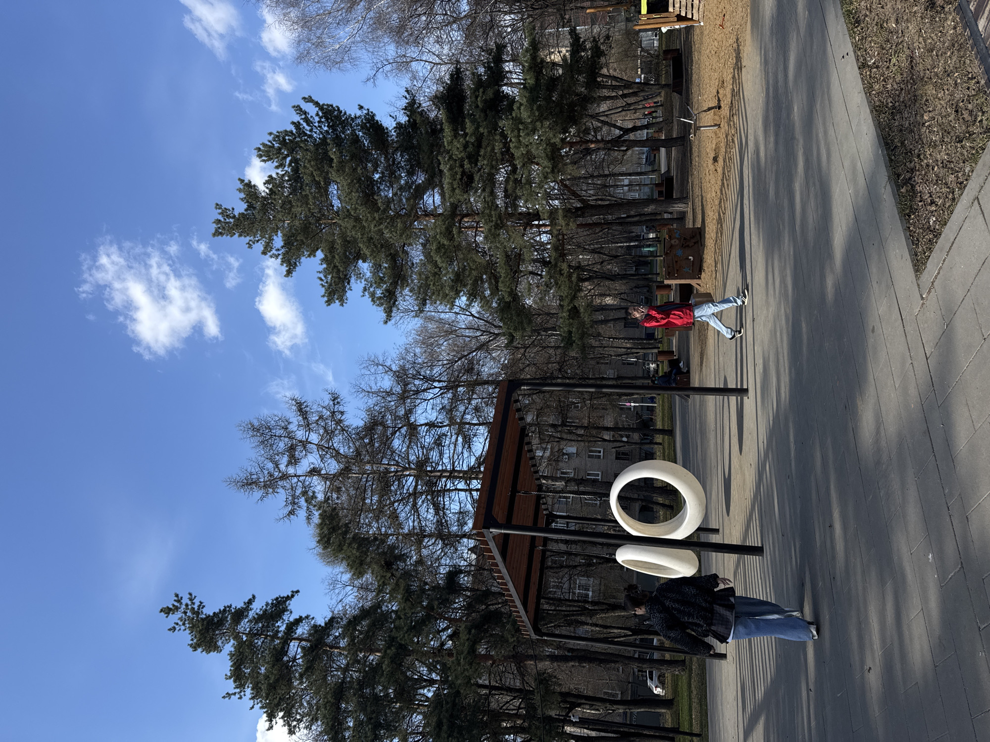 The image shows an outdoor scene in what appears to be a park or public recreational area. Two individuals are present. One person is wearing a red jacket and blue jeans, walking along a paved pathway. A second person, wearing a dark jacket and blue jeans, is walking in the opposite direction. The setting includes a playground area with sand and wooden structures. Tall evergreen trees and other deciduous trees surround the area. Architectural elements, possibly part of the playground equipment, are visible: A structure with a wooden roof and white circular objects suspended. The sky is blue with scattered white clouds. The park appears to be located in an urban area with apartment buildings in the background. This is the city of Moscow, Russia.