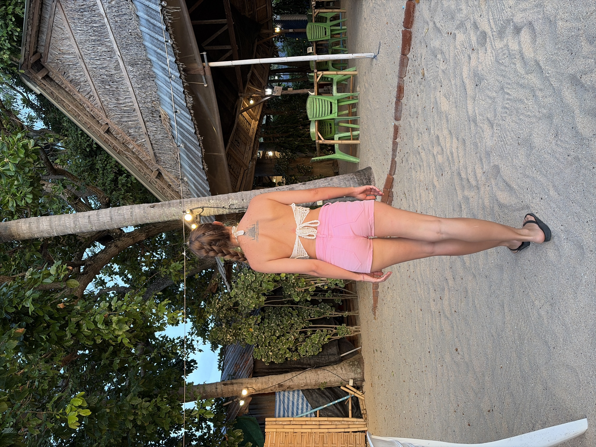 The image depicts a rear view of a woman standing on a sandy beach in Baan Thong Po, Thailand. She is wearing a white crochet halter top and pink shorts. Her hair is in a braid, and she has a tattoo on her upper back. The background shows a rustic structure with a thatched roof, possibly a restaurant or bar, with green plastic chairs and tables visible underneath. Palm trees and lush vegetation surround the area. The ground is covered in sand, with some areas bordered by bricks. String lights are visible, suggesting an evening or early morning setting. The overall atmosphere suggests a tropical, relaxed environment.