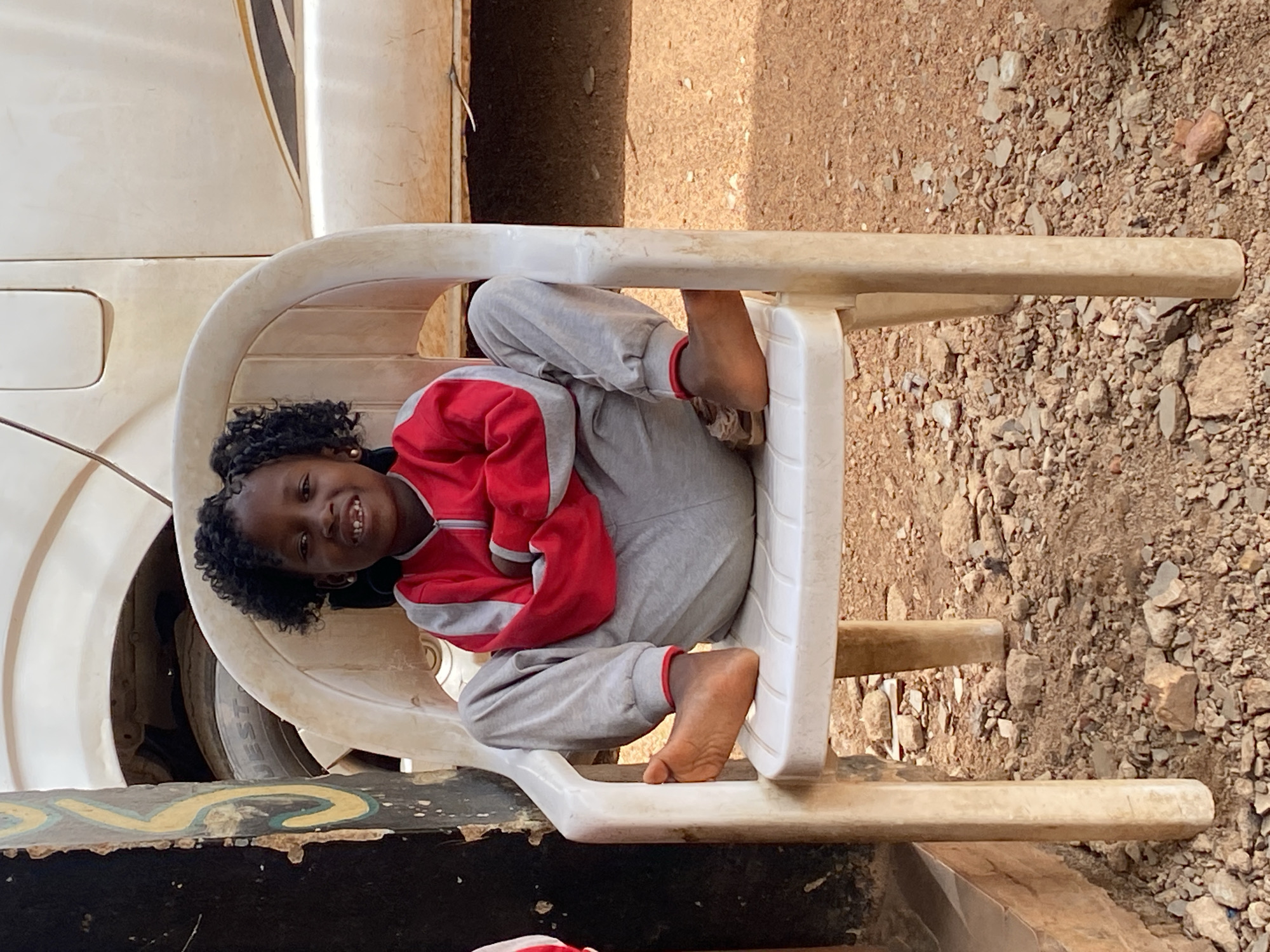 A young girl in Kuru Karama, Nigeria sits contorted in a white plastic chair. She wears a red and gray sweatshirt and gray pants, with bare feet. Part of a white vehicle is visible to the left of the chair. The ground is rocky and dirt-covered.