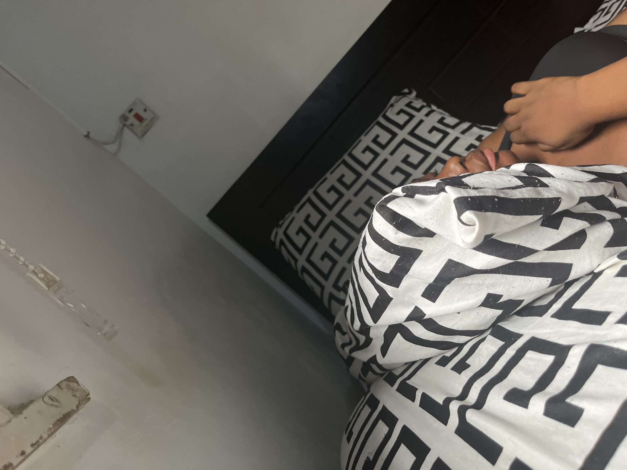 The image shows a person lying in bed with a black and white patterned pillow and blanket. The person's hand is visible. In the background, there is a black headboard and a white wall with an electrical outlet. The location is likely indoors, but the city of Abuja, Nigeria cannot be verified from the image.