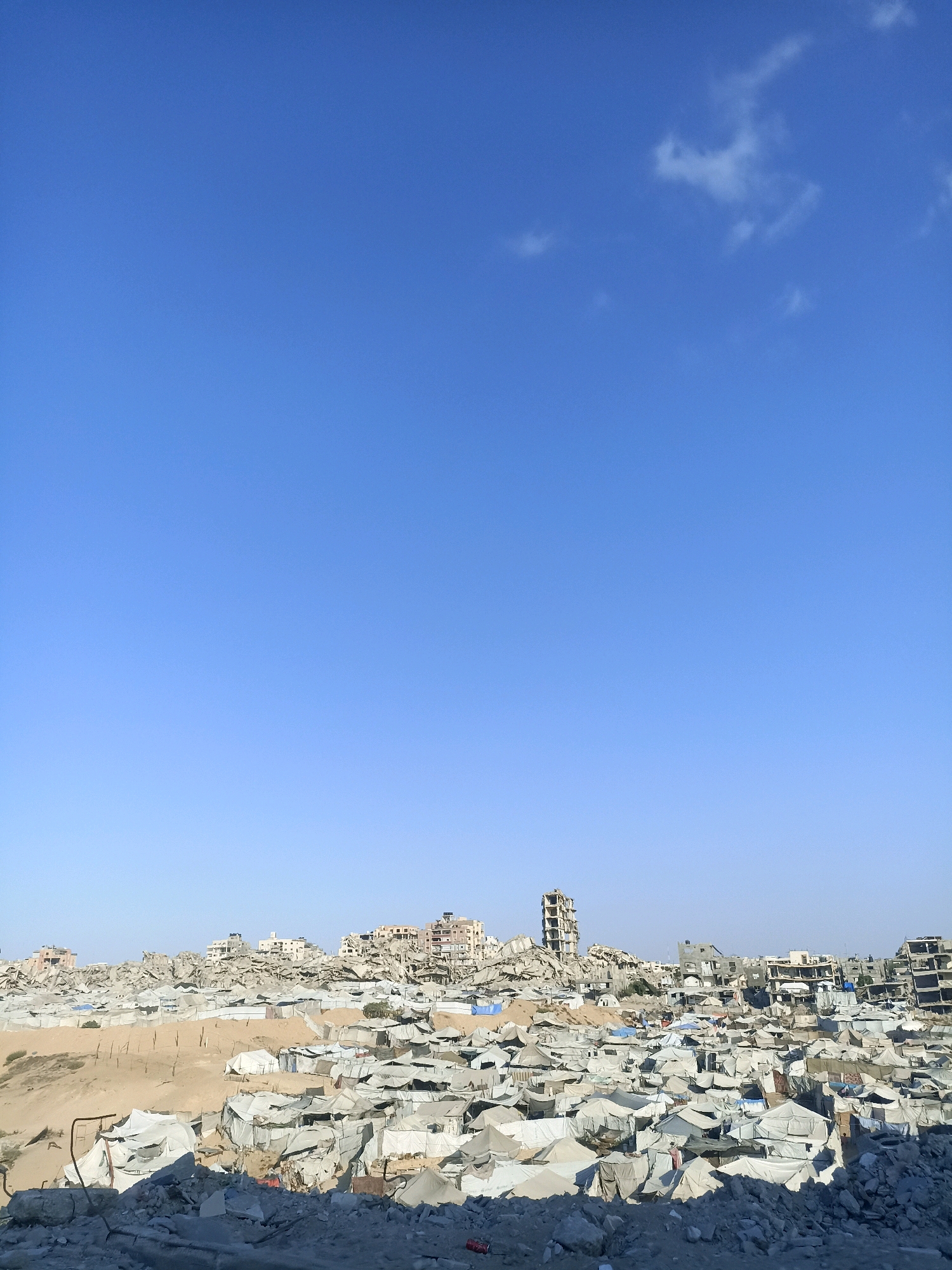 The image shows a wide view of destruction in the Gaza Strip. In the foreground, there are piles of rubble, including large concrete debris. Behind this, a dense settlement of white tents and temporary structures covers a large area, suggesting a displacement camp or makeshift housing. Further in the background, damaged buildings, some partially collapsed and others appearing intact but surrounded by debris, dominate the skyline. The architecture of the damaged buildings indicates a mix of multi-story residential or commercial structures. The sky is clear and blue with a few wispy clouds. The overall scene depicts a landscape heavily impacted by conflict and destruction.
