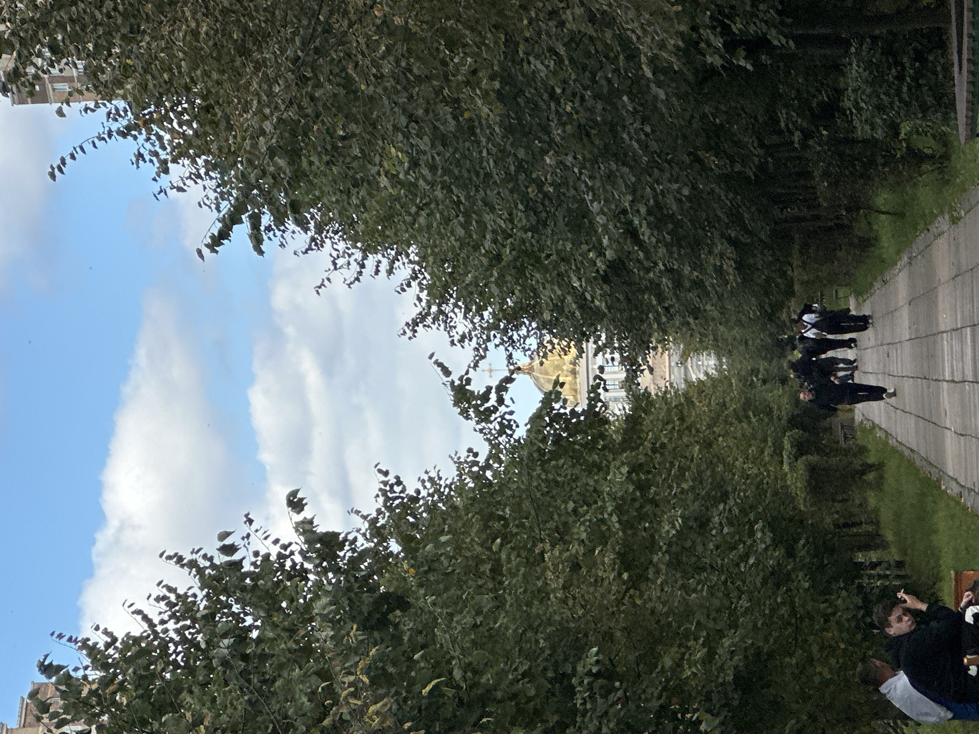 The image captures a pathway lined with trees in Podolsk, Russia. In the background, a building with a golden dome is visible beneath a sky with scattered clouds. Several people are walking along the pathway, appearing to be engaged in casual strolls. In the foreground, two individuals are seated on a bench to the left, with one appearing to be smoking a cigarette. The overall scene suggests a public park or a pedestrian area within the city.