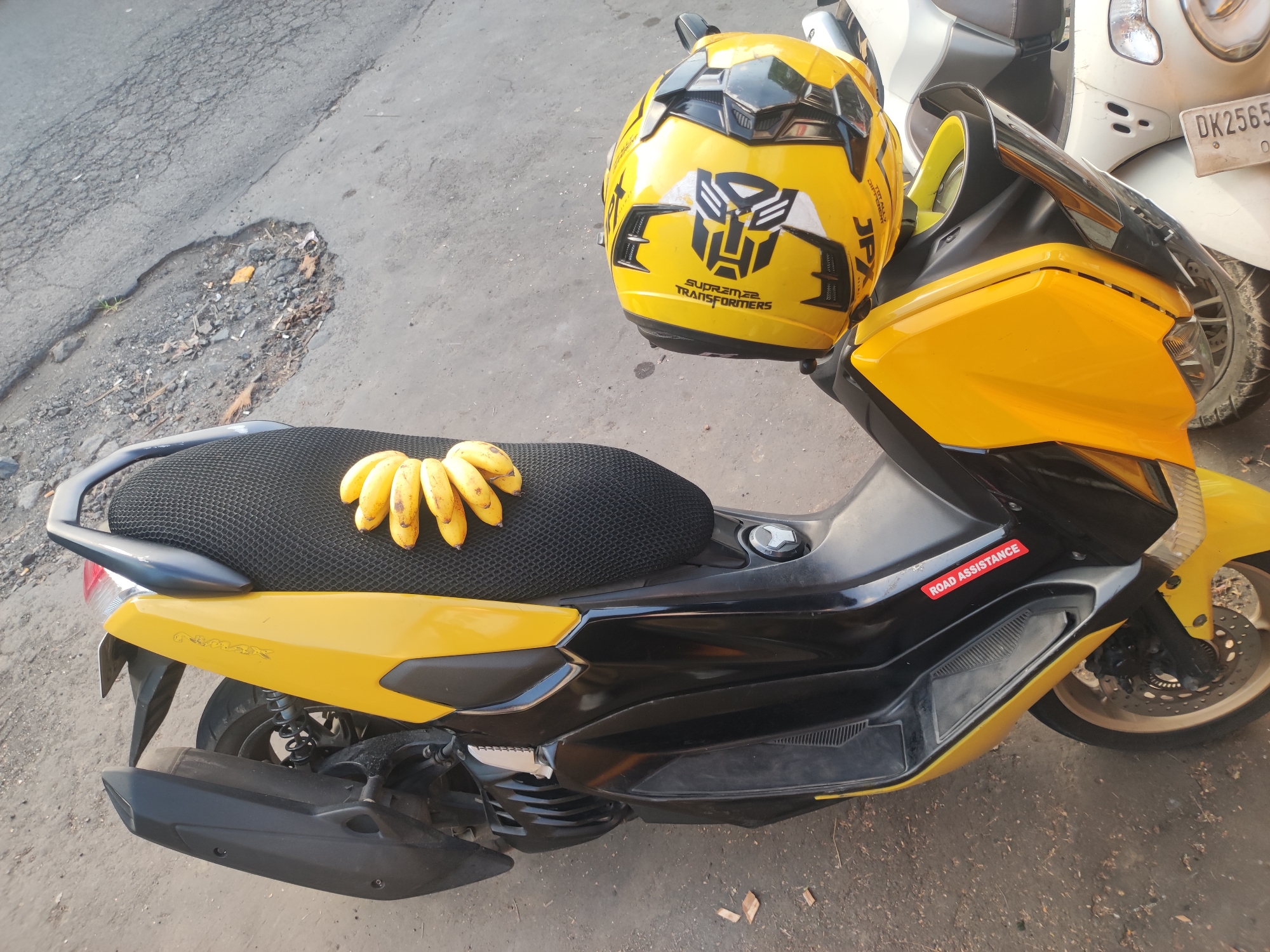 A yellow and black Yamaha NMAX scooter is parked on a paved street, its seat covered with a black mesh pad upon which a bunch of bananas rests. A yellow helmet with a Transformers logo sits on the scooter’s handlebars. The scooter has a sticker that reads "ROAD ASSISTANCE" on its side. To the right, part of another scooter, possibly a white Honda Scoopy, is visible. The environment appears to be an urban area, given the paved road and parked scooters, and is located in the city of Ubud District, Indonesia. The photo is taken from a slightly elevated angle, providing a clear view of the scooter and its immediate surroundings.