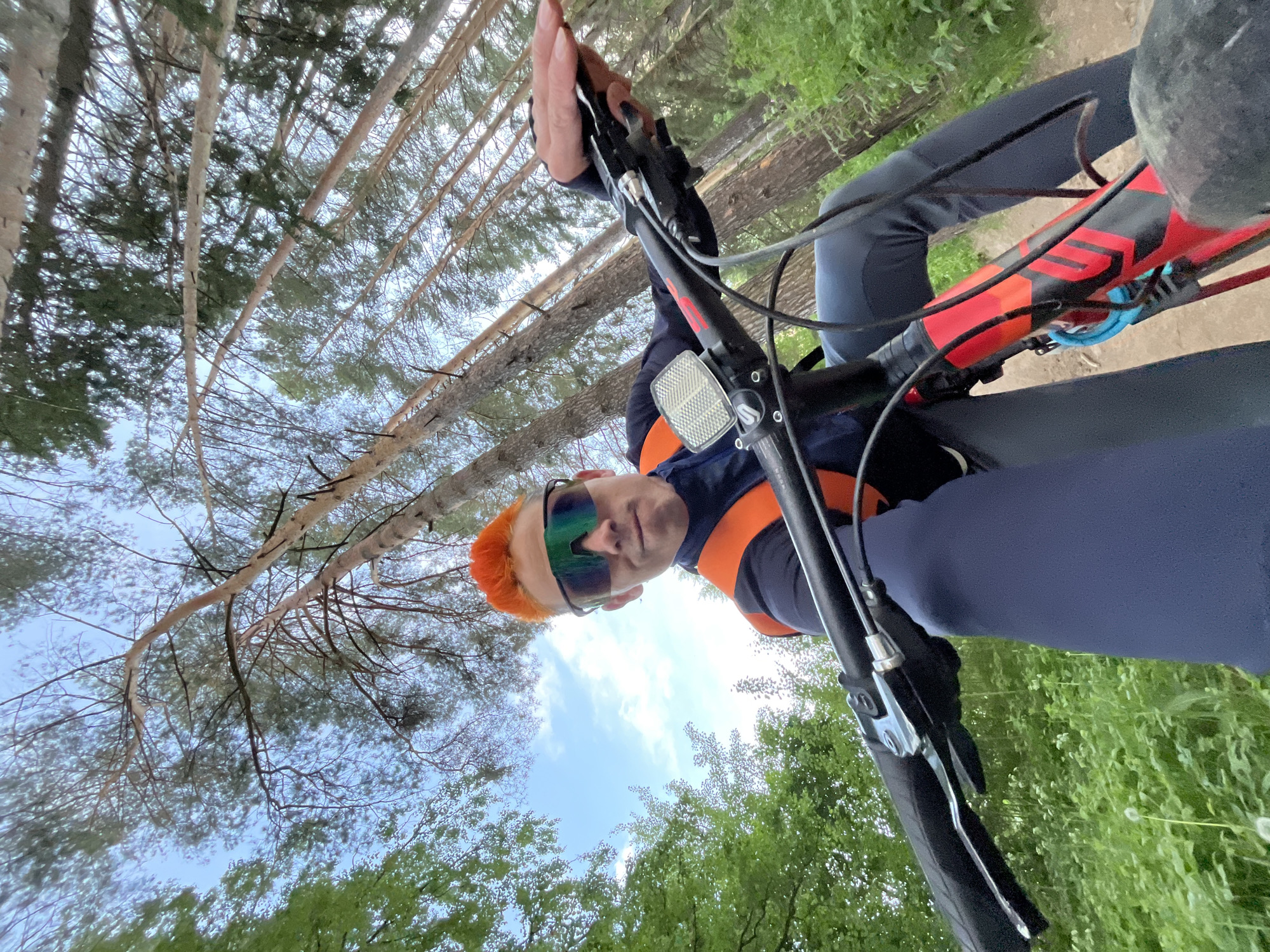 A low-angle, ground-level shot shows a person on a bicycle, looking down at the camera, presumably taken on a trail in a wooded area, likely in Chaikovsky, Russia. The person has dyed orange hair, dark sunglasses, and wears a dark blue top with orange straps or a vest. They grip the handlebars of a bicycle. The bike has a black handlebar, a reflector, and a red and black frame visible. Above, tree trunks and branches reach toward a sky with patches of blue and white clouds, suggesting a forest canopy. Green foliage and trees are seen on the sides of the frame.