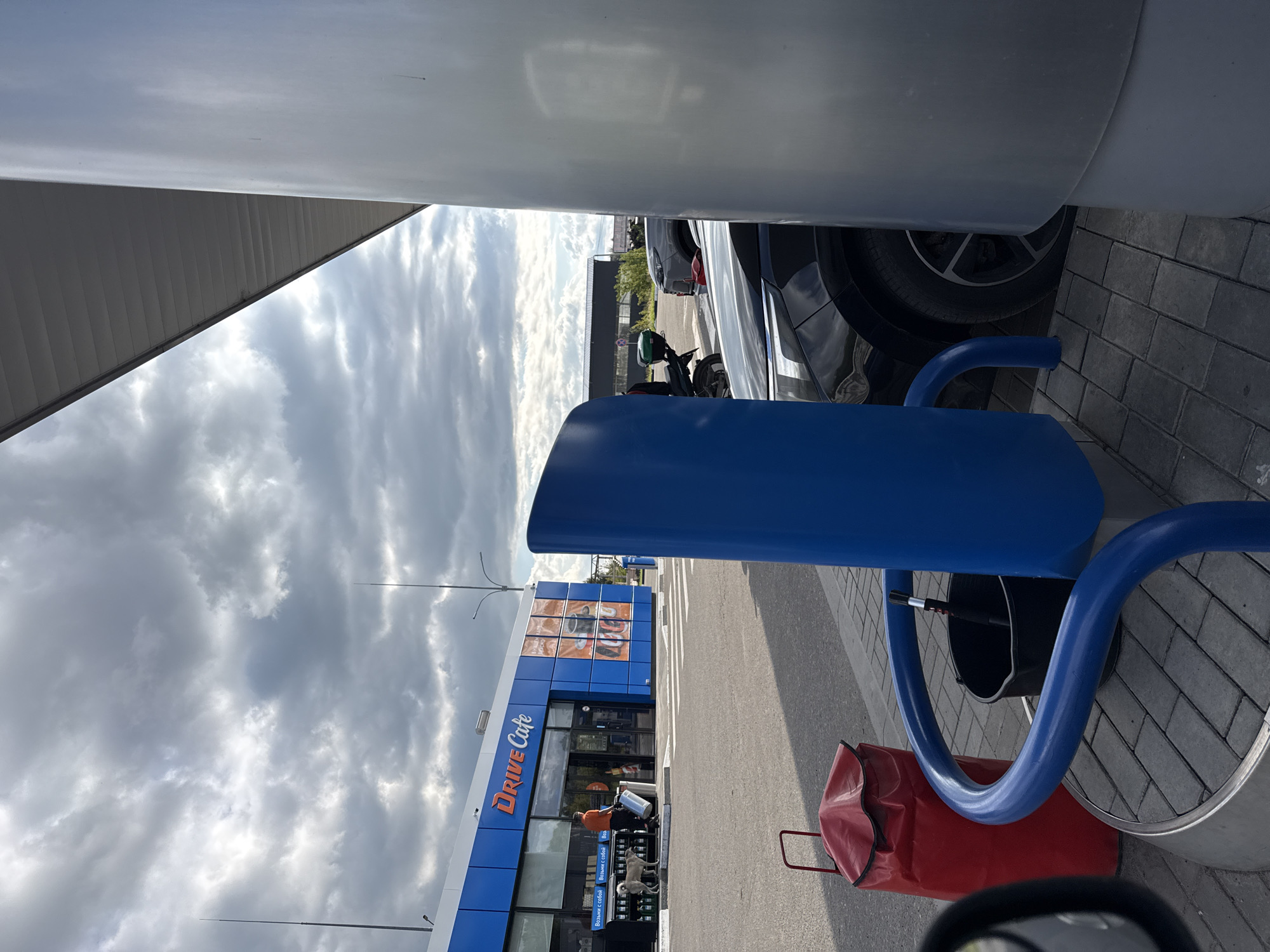 An outdoor scene at a Drive Cafe establishment under a cloudy sky features a blue building with "Drive Cafe" branding. A person in an orange shirt is standing near the entrance with a dog, holding a grey cylindrical object. A red bin with a handle and a black bucket with a cleaning tool are on the paved area. In the foreground, a large, smooth, silver cylindrical pillar obscures part of the view, and a bright blue, curved object is also prominent. Several cars and a motorcycle are parked in the background. The ground is paved with grey interlocking bricks.