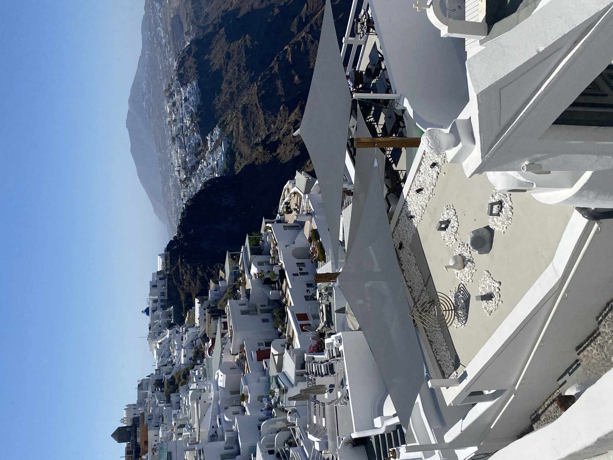 This is a high-angle, eye-level, long shot of a white village clinging to a cliff face in the city of Thira Municipal Unit, Greece, overlooking a caldera and a distant mountain range. The architecture is predominantly white, with cubic structures, flat roofs, and some domed roofs, including at least one blue-domed church. Buildings are densely packed, cascading down the steep slope.
In the foreground, a paved rooftop area is visible, decorated with white pebbles and some decorative objects, including a sculpted bird and a metal artwork. Two large, dark grey shade sails are stretched over parts of this rooftop area, with tables and chairs set up underneath. There are a few people seated at the tables. Another rooftop area, further back, features lounge chairs and umbrellas.
The cliff face is dark and rocky, with sparse vegetation. The sea or caldera water is not directly visible in this framing, but the geological formation suggests its presence. The sky is clear and bright blue. The overall impression is one of a sun-drenched, picturesque Mediterranean resort setting.