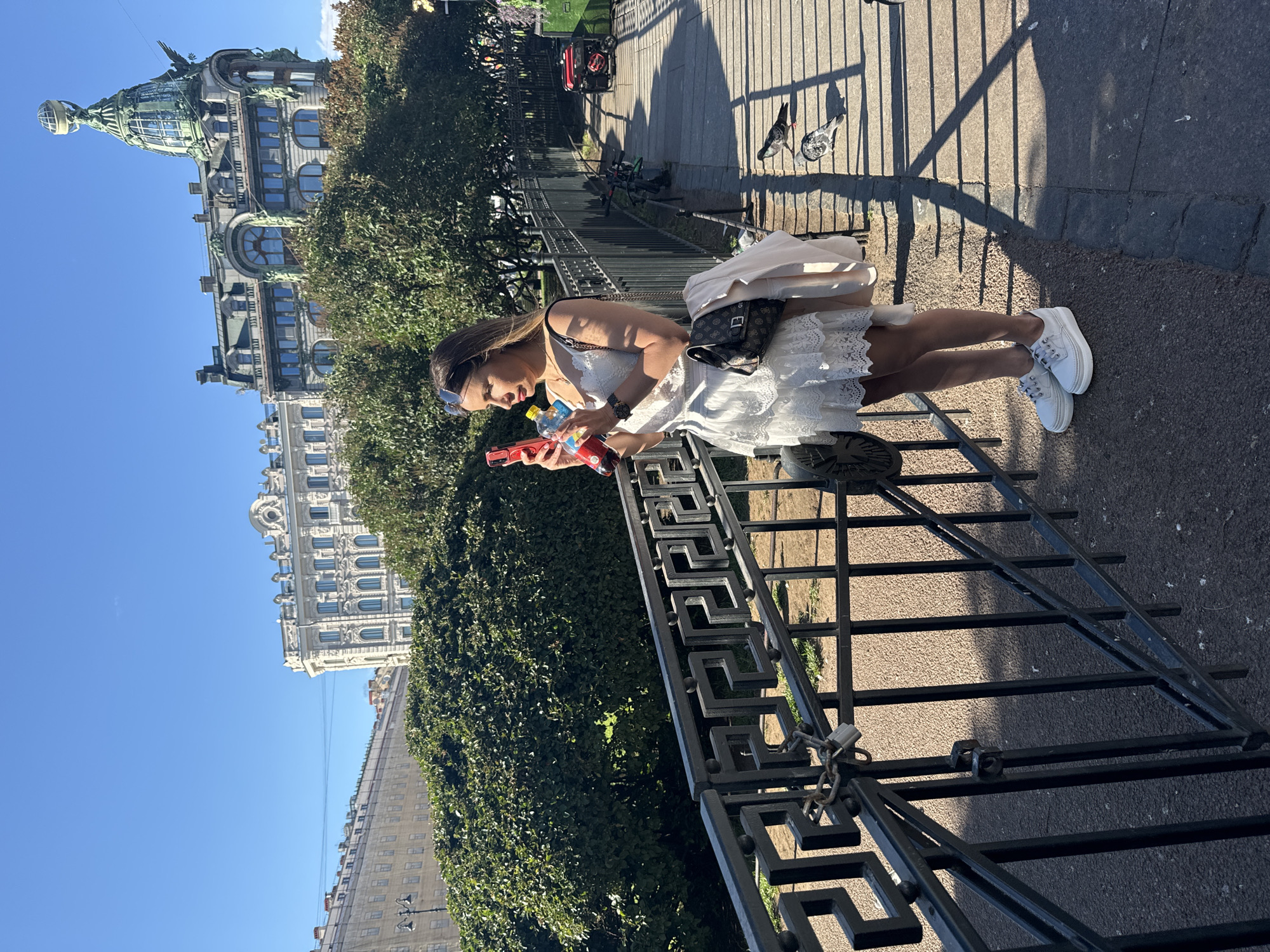 The image shows a young woman in a white lace dress and white sneakers, standing on a paved walkway near a decorative black metal fence. She is holding two bottles, one yellow and one red, and looking down at them. A black fanny pack is worn around her waist. Her expression appears focused. In the background, there are trees and several historic buildings with ornate architecture under a clear blue sky. The sunlight casts long shadows on the walkway, suggesting it is late afternoon. Two pigeons are visible on the walkway near the fence. The overall scene evokes a relaxed, perhaps touristy atmosphere in an urban environment.