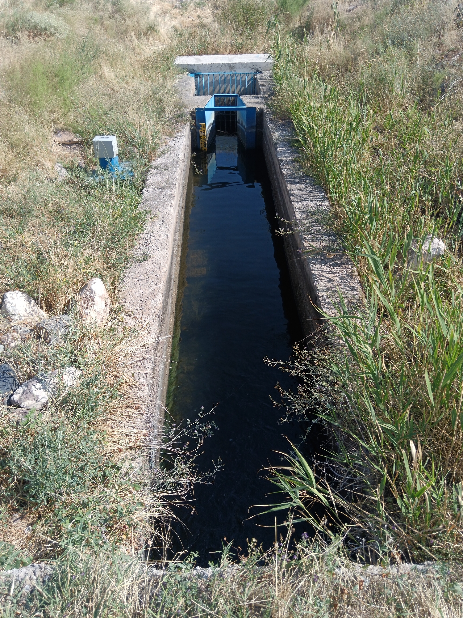 A view from above shows a narrow, dark-water irrigation canal in Aragatsavan, Armenia. The concrete-lined channel runs through dry, grassy terrain dotted with rocks. At the end of the visible canal section, there is a blue metal grate and a structure with a blue railing, suggesting a water control mechanism. A small blue and grey box with an antenna-like protrusion is visible on the left bank of the canal. The surrounding environment is arid with sparse vegetation.