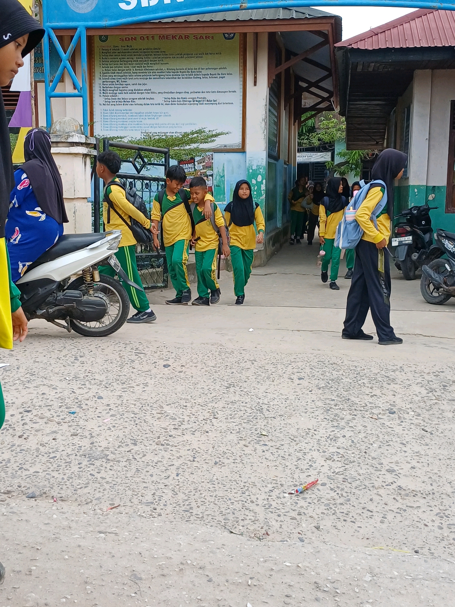 The image shows the exterior of a school building in Dumai City, Indonesia. Several students, mostly wearing yellow shirts and green pants, are walking and interacting outside. Some are carrying backpacks. A motorcycle is parked to the left of the frame. A sign on the building reads "SDN 011 MEKAR SARI". The ground is paved with asphalt and small stones. In the foreground, a small, colorful object, possibly a firecracker, lies on the ground.
