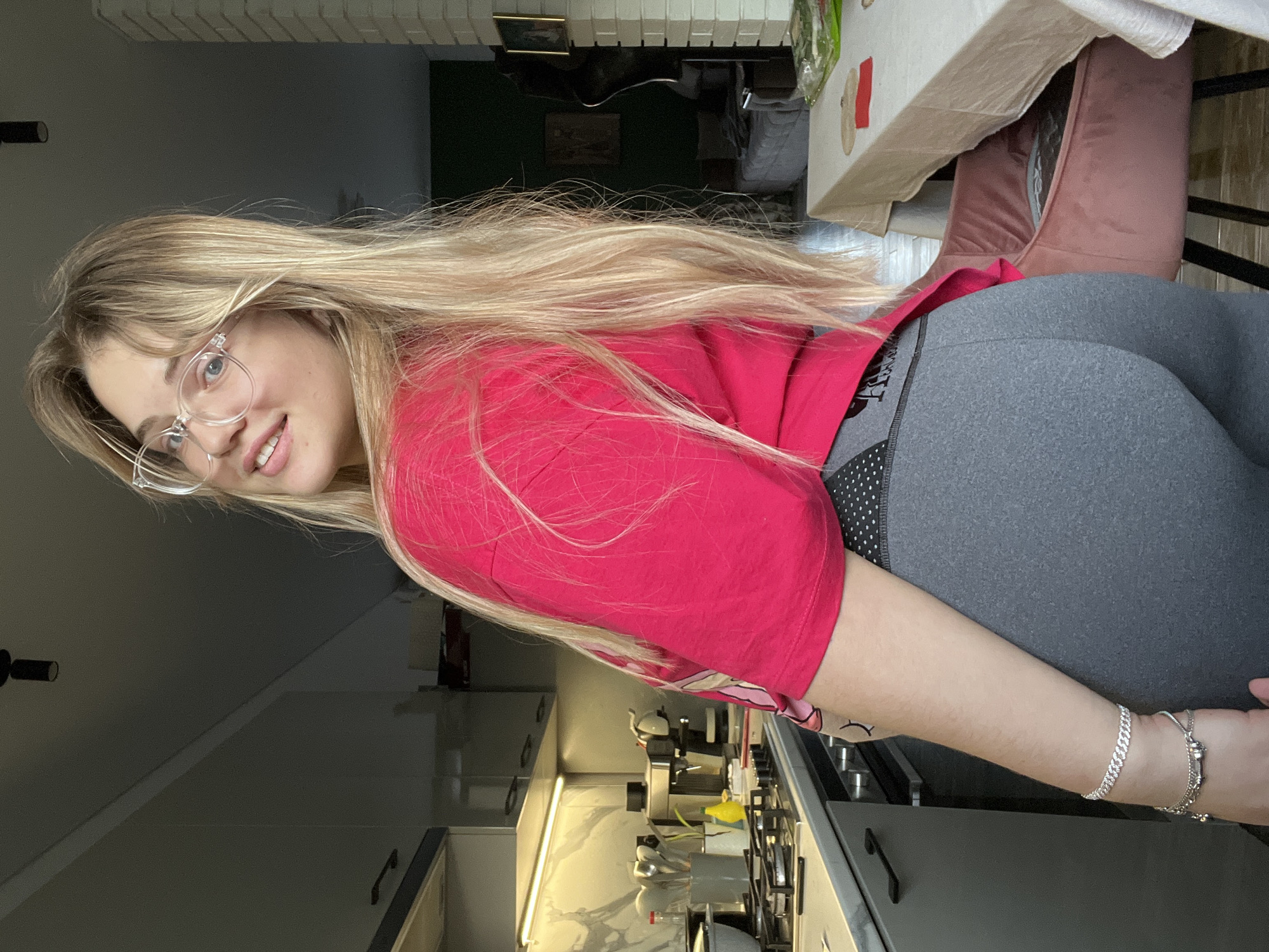 The image captures a young woman posed indoors, possibly within a home in Большая Шильна, Russia. She has long, straight blonde hair and is wearing clear-framed glasses. Her head is tilted, and she looks directly at the camera with a gentle smile.
She is dressed in a vibrant red short-sleeved t-shirt and dark grey, form-fitting leggings. Visible on the waistband of her leggings, partially obscured, is the brand name "PINK." On her left wrist, she wears several silver-colored bracelets. A small, stylized eye tattoo is visible on her left upper arm.
The background suggests a modern home interior, encompassing both a kitchen and a dining area. In what would be the lower part of an upright image, a contemporary kitchen counter with under-cabinet lighting, a stove, and an espresso machine are visible. Above her, on the right side of the frame, is a dining table covered with a light-colored tablecloth, beside which sits a chair with pinkish-brown velvet upholstery. The lighting appears to be natural, consistent with daytime.