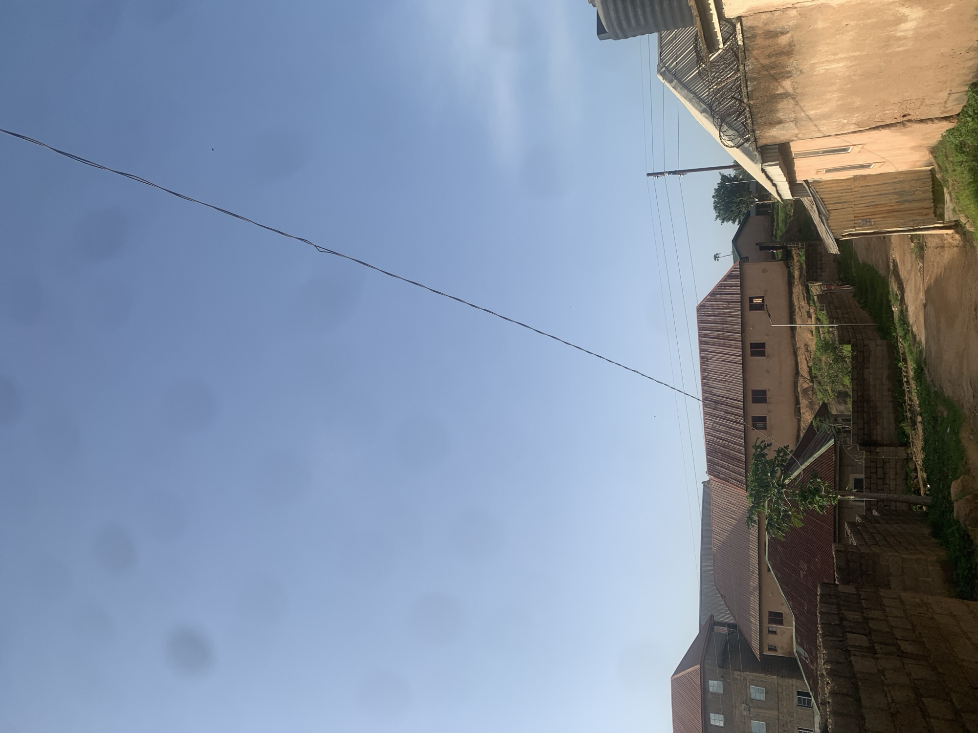 This image depicts an outdoor scene in Jos, Nigeria, under a clear blue sky. The perspective is a low-angle shot, looking upwards. A prominent feature is a thin, dark wire stretching diagonally across the sky.
In the background, the scene is dominated by buildings with predominantly corrugated metal roofs in shades of red and brown. One building appears to be a multi-story structure with visible windows. Another building, closer to the foreground, has a peach-colored exterior wall and a corrugated roof. There are also sections of brick walls and a patch of green grass visible. A small tree with green foliage grows in front of one of the buildings.
The time of day appears to be daytime, suggested by the bright sky. The weather is clear with no clouds visible. The location cues of corrugated roofs and the general architectural style suggest a residential or mixed-use area in Nigeria. There is no visible text in the image. No people or specific activities are clearly depicted. The overall impression is one of a daytime view of an urban or semi-urban environment in Jos, Nigeria, characterized by its buildings and the sky above.