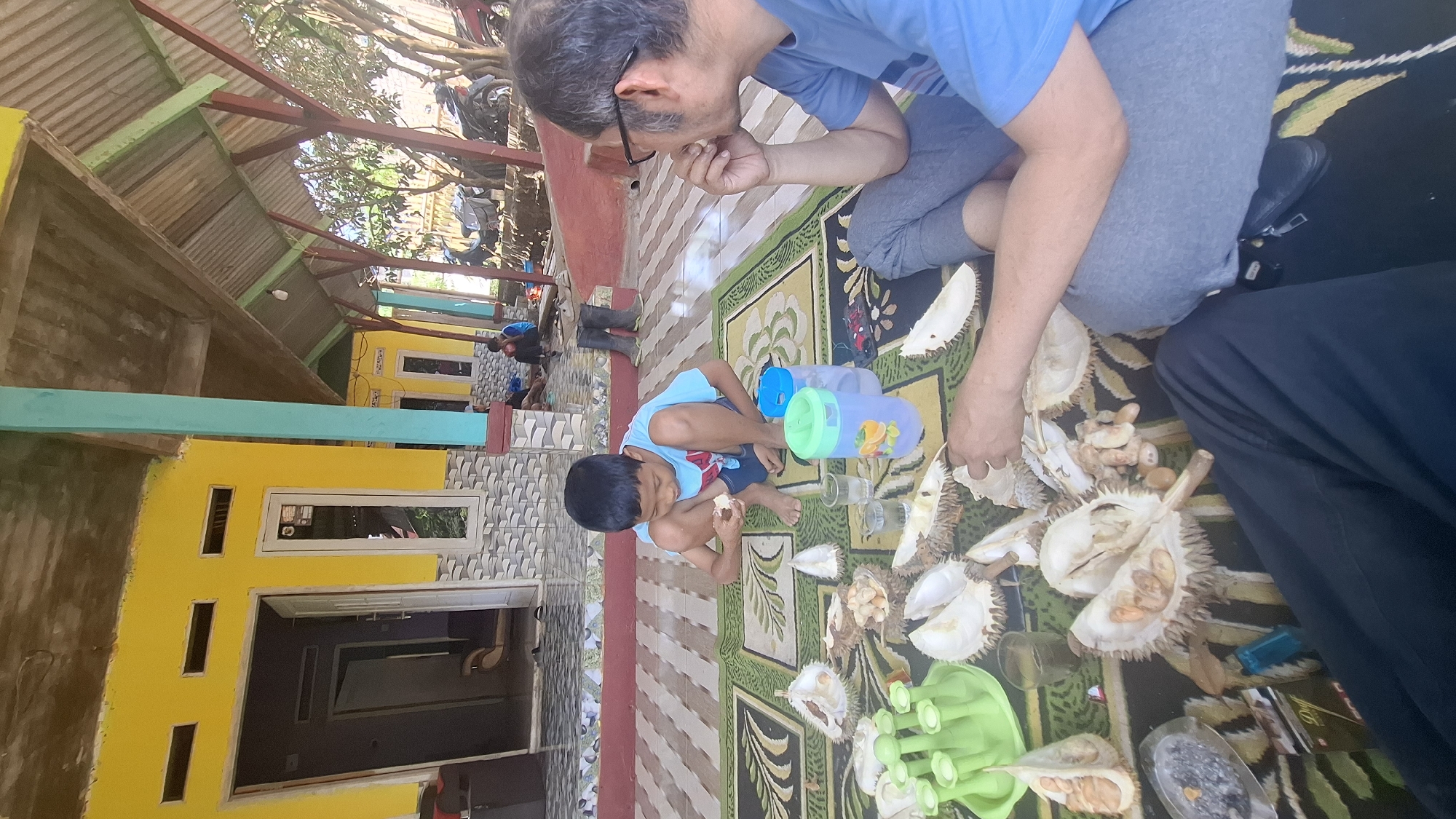In Cilaku, Indonesia, a young boy in a blue t-shirt and shorts sits on a patterned rug, eating durian. A man with glasses, wearing a blue t-shirt and gray pants, sits to the boy's right, looking away from the camera. Scattered around them are numerous opened durian fruits, their spiky husks and creamy flesh visible. A green durian holder, several glasses of water, and a bottle with a green lid are also present on the rug. In the background, a yellow house with a blue trim stands under a covered patio area. Two other individuals, possibly children, are visible sitting further in the background, near the entrance of the house. The environment appears to be an outdoor, covered seating area attached to a residential building, with motorcycles and trees visible in the distance. The lighting suggests it is daytime.