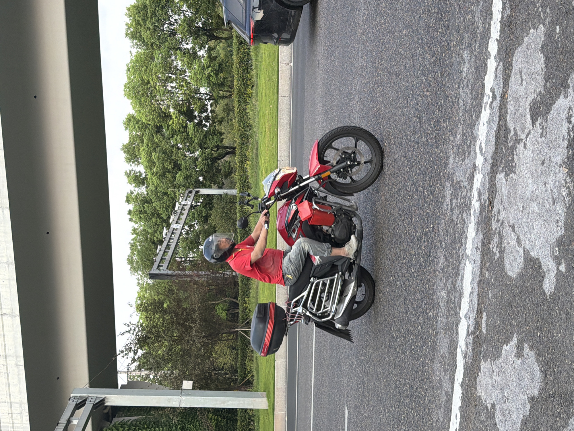 A man is riding a red motorcycle with a black storage box on the back. He is wearing a red shirt, camouflage pants, and a black helmet with a clear visor. The motorcycle has a silver rack on the back and a silver fender. The man is on a paved road with a white line marking the lane. In the background, there are trees and a gray overhead structure. A black SUV is visible on the right side of the image. The image appears to be taken on a cloudy day. The location is identified as Jiangjin, China.