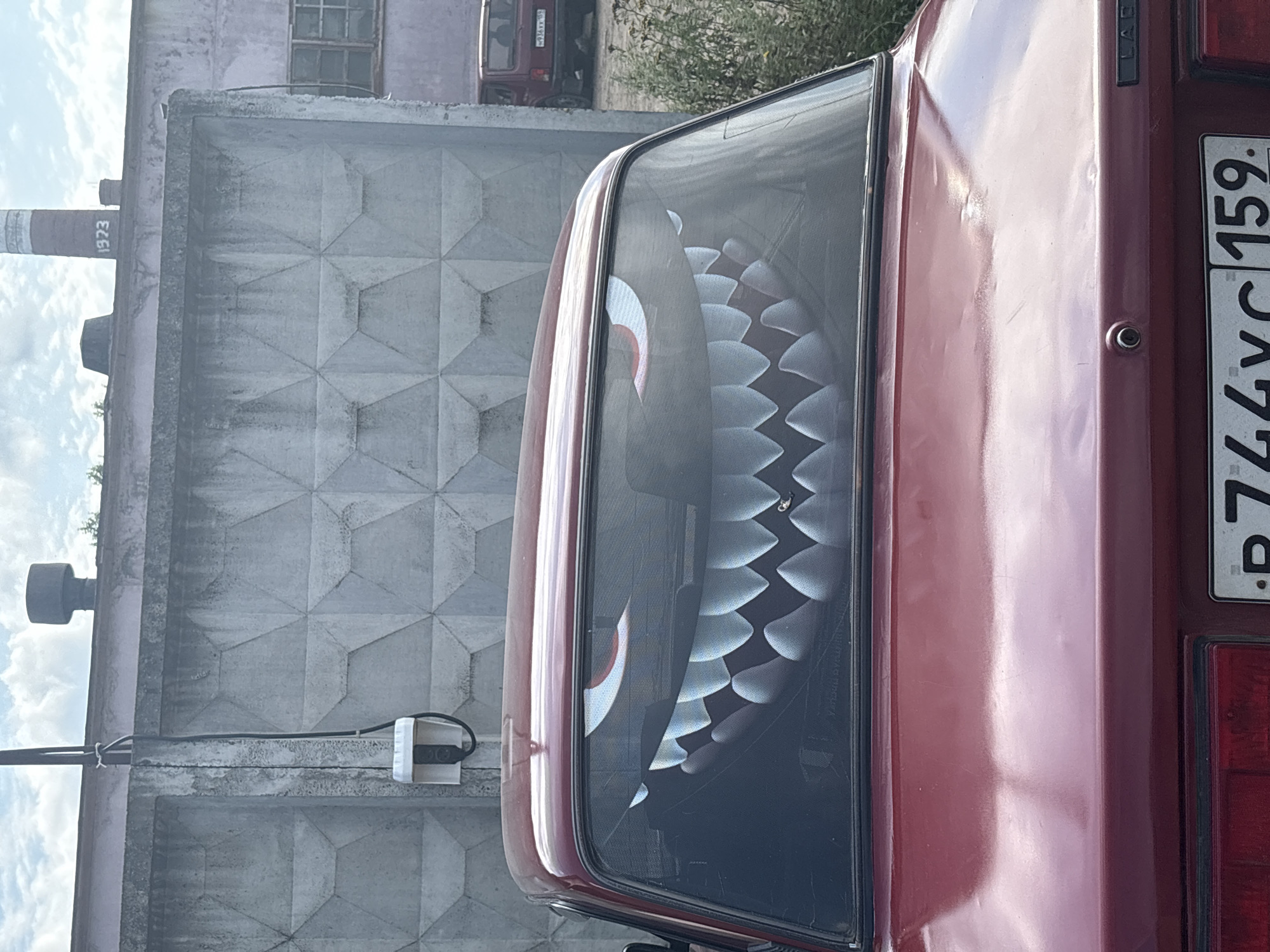 The image captures a close-up rear view of a dark red sedan, likely a Lada, parked outdoors in front of a textured concrete wall. The car's rear window features a prominent decal of a cartoonish, monstrous mouth with sharp, white teeth and red-outlined eyes, giving the impression of a snarling face. A single, small dark object, possibly a piece of debris, is visible on the lower right portion of the decal. The car's trunk is slightly dented on the right side, reflecting the sunlight.
Behind the car, the concrete wall is made of interlocking diamond-shaped blocks. To the left of the car, a black electrical cable runs vertically down the wall, attached to a white electrical box. In the background, beyond the wall, a red building with windows is visible, and further back, a tall, red-and-white striped industrial chimney with the number "1973" painted on it stands against a cloudy sky. Another, smaller chimney is to its left. To the right of the car, a bushy, overgrown plant with dark green leaves is present. The overall setting suggests an industrial or urban environment in the city of Noviy, Russia.