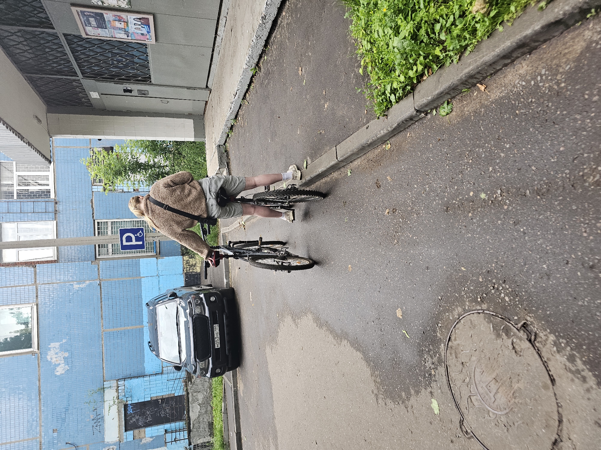 A person with blonde hair, wearing a fuzzy brown jacket, grey shorts, white socks, and beige sneakers, stands on a street in Moscow, Russia, looking at their phone. They are positioned between two bicycles, one in front of them and one behind, both appearing to be on a paved surface next to a curb. A black SUV is parked to the left of the person and bicycles, with its front facing the viewer. The background features a blue apartment building with visible windows and graffiti on the lower left side, as well as a gray building entrance to the right. A blue parking sign with a white 'P' is visible on a pole near the apartment building. The ground is wet, suggesting recent rain. A circular manhole cover is visible on the wet pavement in the foreground.