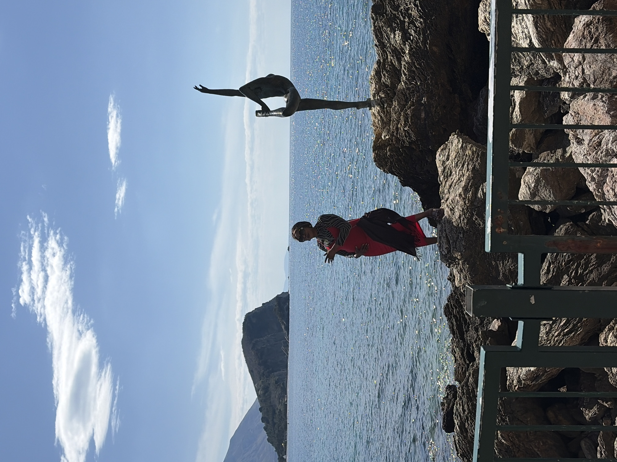 The image shows a coastline on a sunny day. The statue of a male figure doing yoga sits on a rocky outcrop overlooking the sea. A woman in a red dress stands on the rocks near a metal railing. In the background is the coastline, with a large mountain in the distance. The location is Budva, Montenegro, specifically the stretch of coastline at the end of the promenade close to the Budva Old Town. The water is sparkling with sunlight.