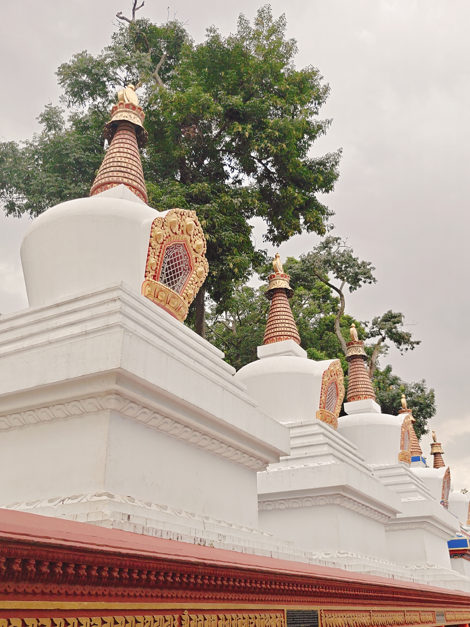 A photograph captures a side view of a row of white stupas with ornate golden detailing, set against a backdrop of lush green trees and an overcast sky. The stupas are arranged in a line, receding into the distance and suggesting a spiritual or monastic site. Each stupa features a rounded dome topped with a tiered, conical spire decorated with bands of red and gold, culminating in a golden finial. Intricate golden architectural elements, resembling windows or decorative panels, are embedded on the sides of some stupas. The foreground reveals the textured red and gold decorative elements of a low wall or railing, with what appears to be decorative script or patterns. The image is likely taken in Kathmandu Metropolitan City, Nepal, a region known for its numerous Buddhist stupas and monasteries. No people or animals are visible in the frame, and the overall atmosphere is tranquil and serene.
