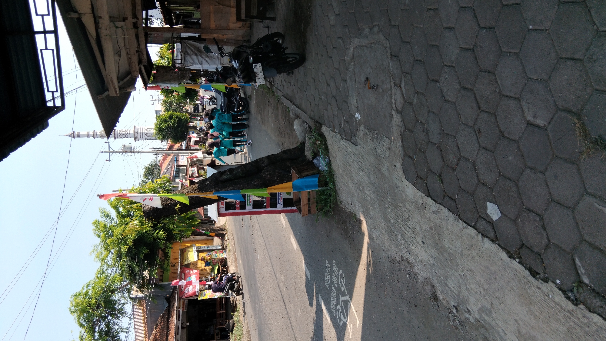 This image depicts a street scene in Proyonanggan Tengah, Indonesia, on a clear day. The foreground shows a paved sidewalk with hexagonal stones, adjacent to a road marked with a bicycle lane symbol and the Indonesian text "JALUR KHUSUS SEPEDA" (Special Bicycle Lane). To the left of the road, there are several buildings with tiled roofs and shops.
Along the sidewalk, a large tree is positioned next to a red and white cylindrical structure adorned with colorful flags. Further down the street, a group of young people wearing teal uniforms are walking together. In the background, a white minaret of a mosque is visible against the sky. A motorcycle is parked on the sidewalk to the right, and another motorcycle is seen on the road in the middle distance. A person on a motorcycle is also seen riding on the road. The overall environment suggests a typical urban street in Indonesia, with a mix of commercial buildings, residential areas, and public infrastructure.