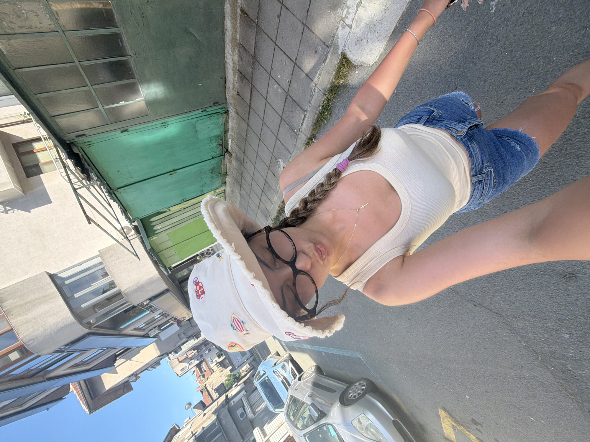 The image shows a young woman standing on a street in Burgas, Bulgaria. She is wearing a white bucket hat with fish patterns, glasses, a cream-colored tank top, and denim shorts. Her hair is braided and tied with a purple elastic band. She is making a kissing gesture with her lips pursed. In the background, there are buildings, cars parked on the street, and a sidewalk. The architecture suggests a European city setting. The street appears to be paved with asphalt, and there are some yellow markings on it. The overall lighting suggests it is daytime, likely in the afternoon due to the shadows. The presence of cars and the urban infrastructure confirm it is a city environment.