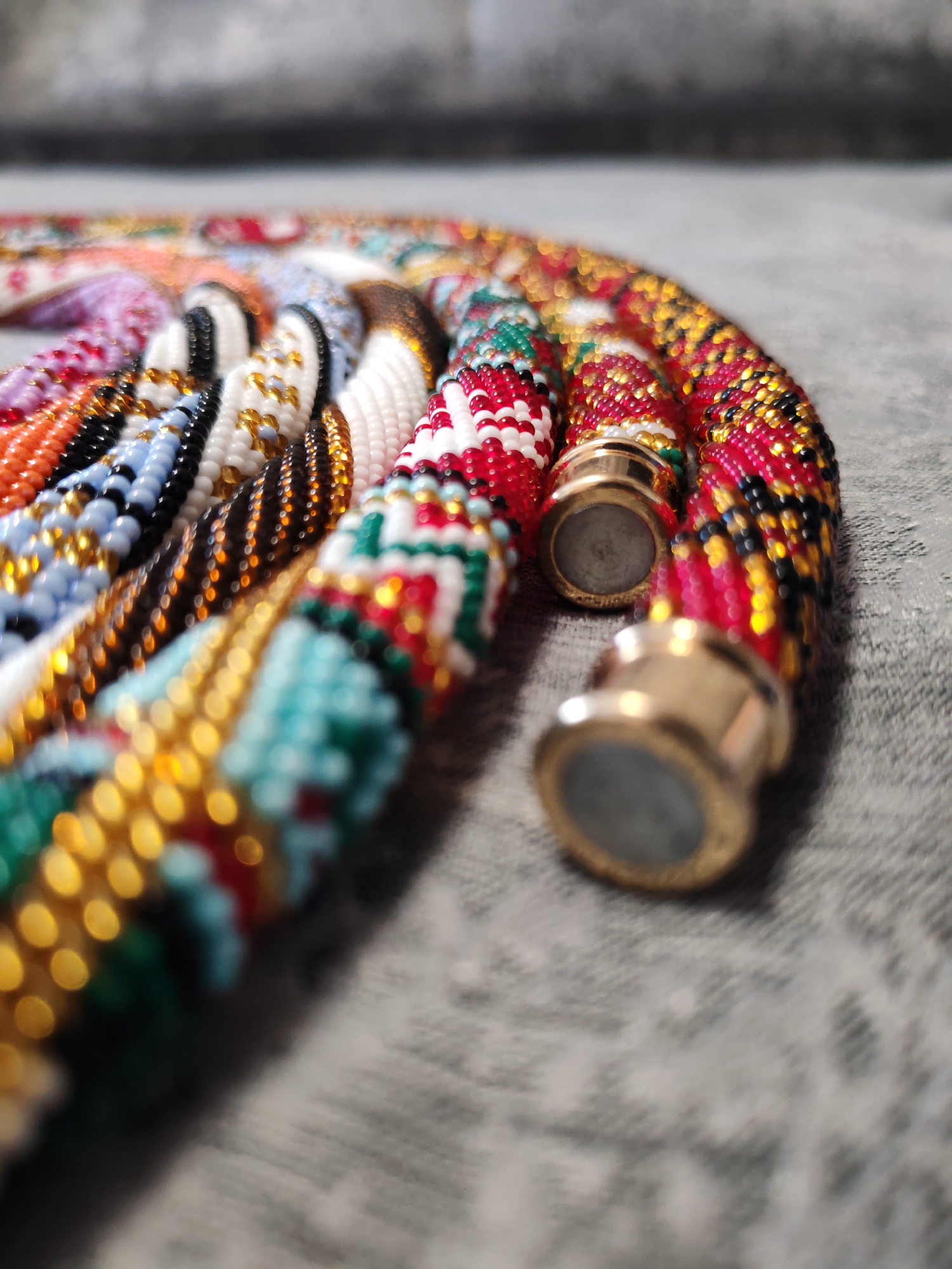 The image shows a close-up, angled view of several beaded necklaces or cords lying on a textured grey surface. The beads are small and densely packed, forming intricate patterns in various colors, including red, white, black, gold, blue, orange, and green. Each beaded cord appears to have a metallic clasp at its end, which are gold-colored and appear to be magnetic. The background is softly blurred, suggesting a shallow depth of field. The lighting is warm and highlights the textures and shine of the beads and clasps. The location is Unknown, Unknown.