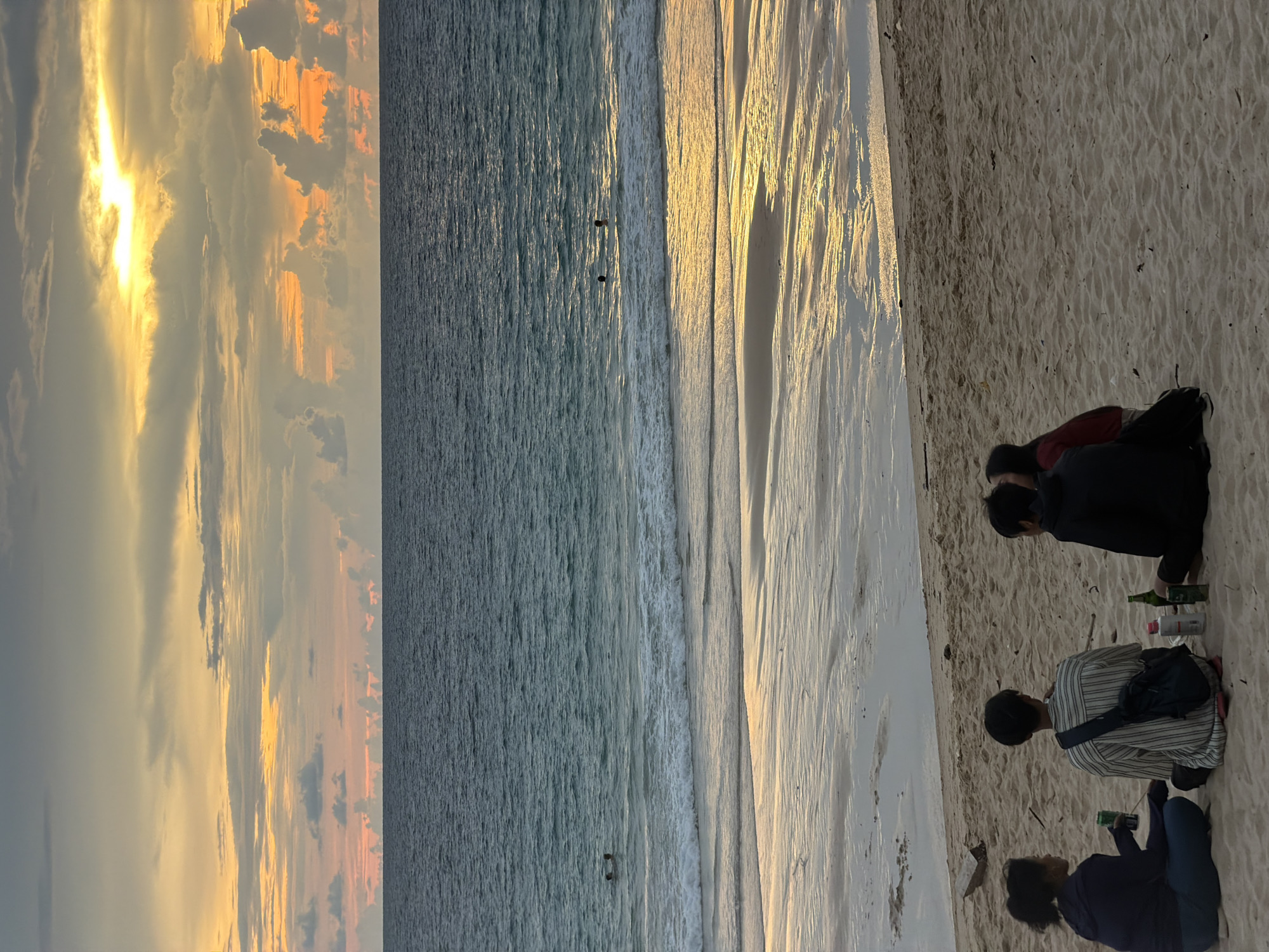 The image depicts a serene beach scene at sunset in Choeng Thale, Thailand. The sky is a canvas of warm colors, with the sun breaking through dramatic clouds, casting a golden glow on the water and sand. The ocean is calm, with gentle waves lapping at the shore, reflecting the vibrant sky.
Several people are present, enjoying the tranquil evening. Two individuals are visible in the shallow water, appearing to be wading or swimming. On the sandy beach, a small group is gathered. One person is seated on the sand, facing away from the camera, with a backpack. Another person is sitting in a striped shirt, holding a drink. A third individual, wearing a dark top and blue pants, is also seated on the sand, holding a green bottle, possibly part of a casual gathering.
Notable details include the striking colors of the sunset, the reflections on the wet sand, and the presence of a few bottles, suggesting a relaxed social occasion. The overall mood is peaceful and contemplative, amplified by the natural beauty of the setting sun over the ocean. The time of day is clearly sunset, and the weather appears to be clear with some dramatic cloud formations. The location is identifiable as a beach, likely in a tropical region given the vegetation implied by the general context of Thailand. No visible text is present in the image.