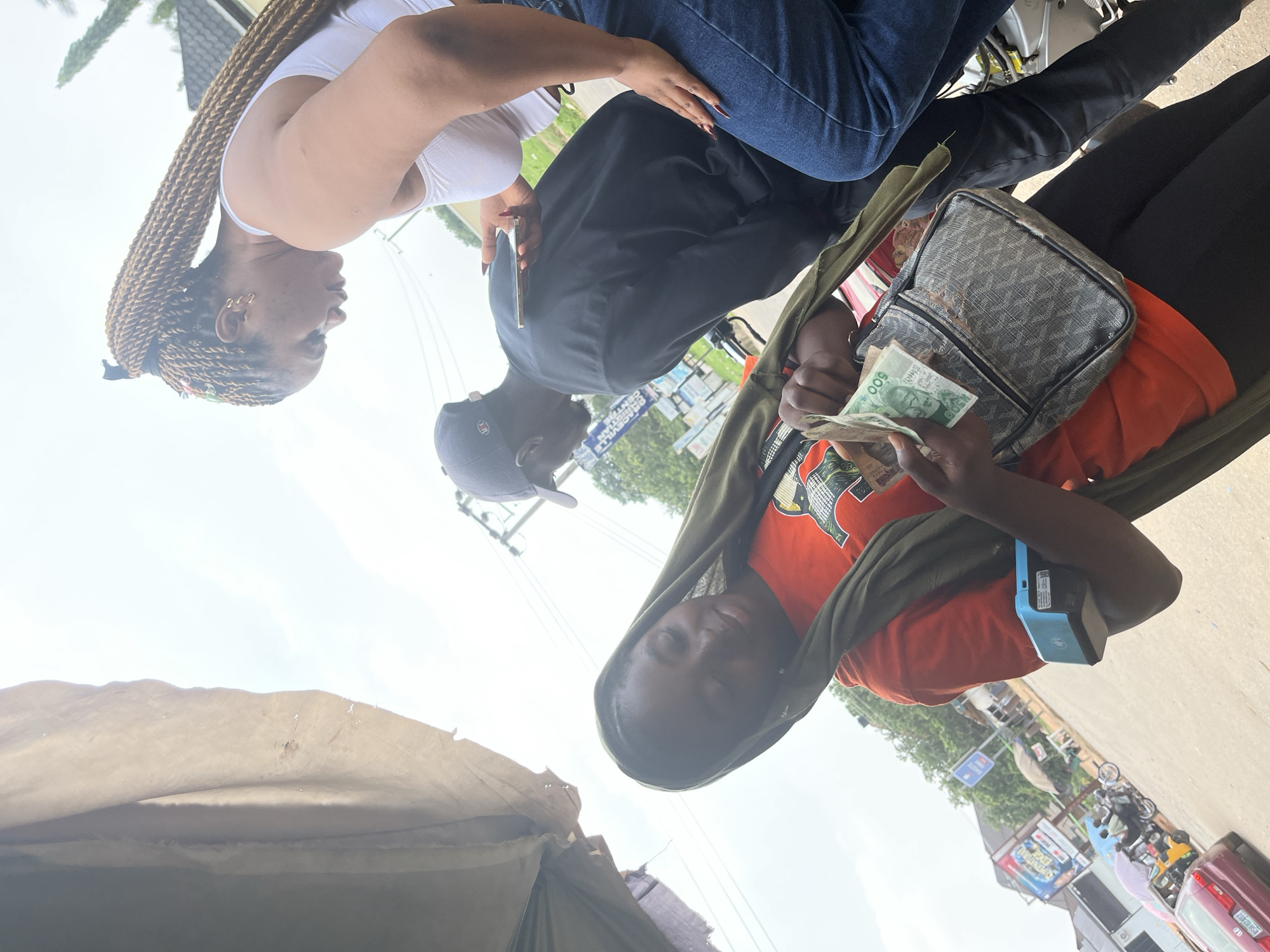 In an outdoor street scene in Abuja, Nigeria, three individuals are present. A young woman in the foreground, wearing an orange t-shirt and a green headscarf, is holding Nigerian Naira currency in her right hand and looking down at it. A portable point-of-sale device is attached to her left arm. Beside her, a woman with long braided hair, wearing a white crop top and blue jeans, is looking at a mobile phone she holds in her left hand. Behind them, a man wearing a black t-shirt and a baseball cap is also looking at his phone. The background shows a street with a few vehicles, buildings, and utility poles. The lighting suggests it is daytime.