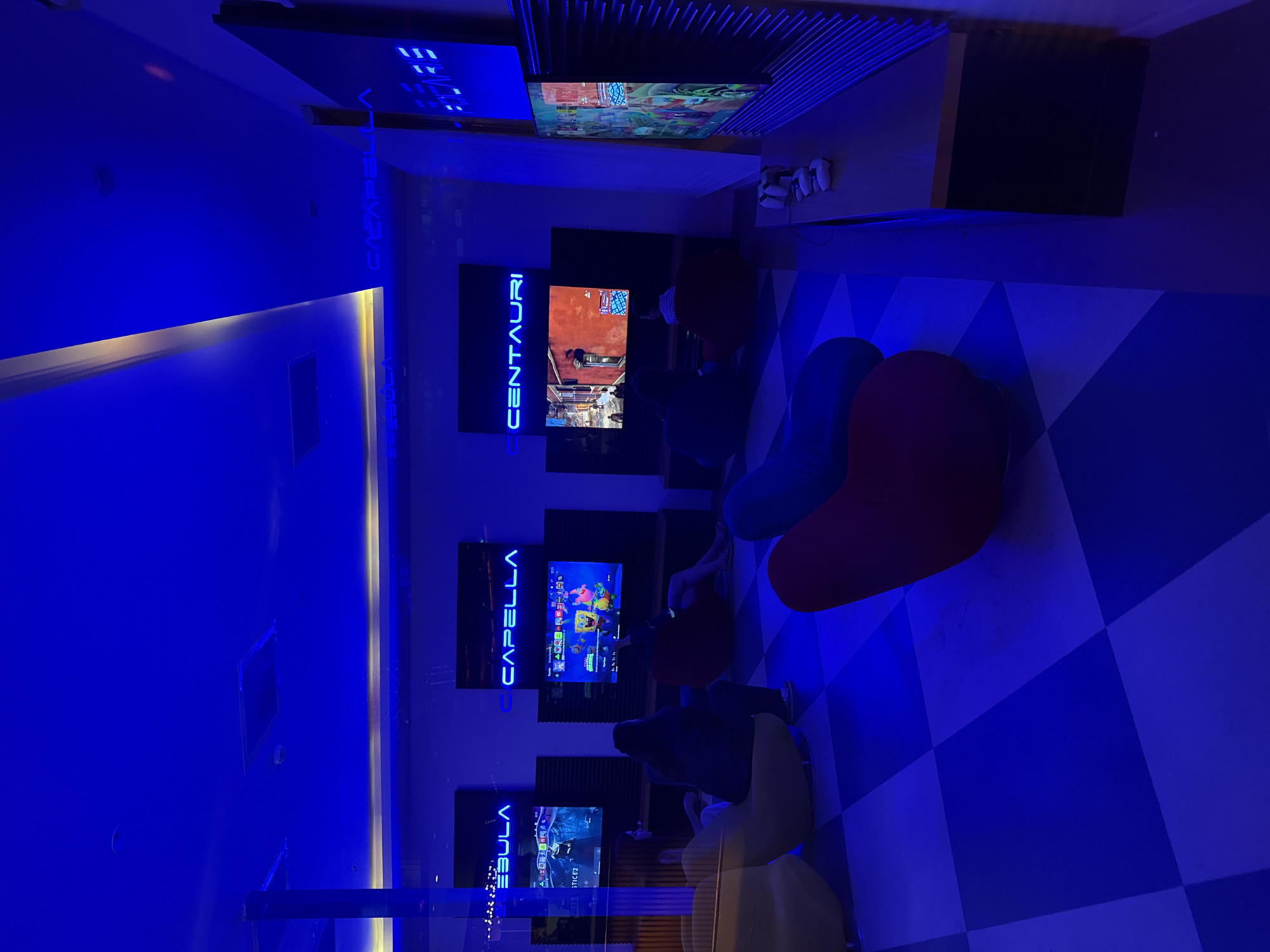 The image depicts an indoor entertainment area, possibly a gaming lounge, illuminated by vibrant blue neon lighting. Several large screens display video games. In the foreground, beanbag chairs in red, blue, and yellow are arranged on a checkered floor. Some individuals are seated in the chairs, facing the screens. In the background, more screens and gaming setups are visible, along with signage that appears to read "NEBULA," "CAPELLA," and "CENTAURI." The overall atmosphere suggests a modern, high-tech entertainment space. The location is identified as Bodrum, Turkey.