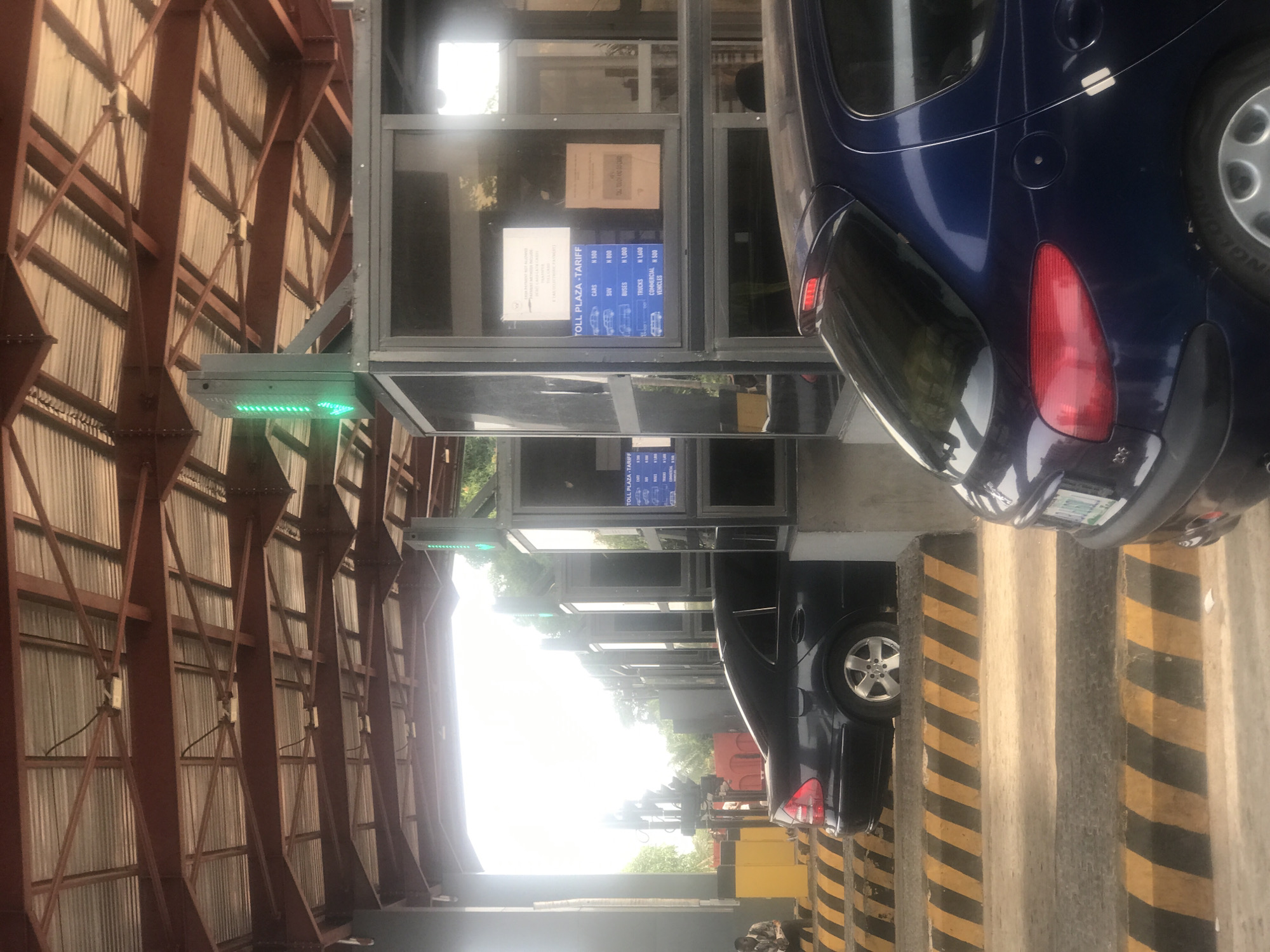 The image captures a bustling toll plaza in Kubare, Nigeria, during bright daylight. Vehicles are actively passing through multiple lanes, with a green arrow illuminated on a traffic light indicating an open lane.
In the foreground, a dark blue car, possibly a minivan or SUV, is prominently featured, with its rear and side visible as it proceeds through a toll lane. Another dark vehicle can be seen further down an adjacent lane. To the left of the vehicle lanes are several glass-enclosed toll booths. One booth clearly displays a "TOLL PLAZA-TARIFF" sign on its window. The overhead structure is a large, covered area supported by reddish-brown metal beams and corrugated metal siding, extending across the toll lanes. The ground features distinctive yellow and black striped markings.
Notable visible text from the toll tariff sign includes:
* "TOLL PLAZA-TARIFF"
* "CARS N 500"
* "SUV N 800"
* "BUSES N 1,000"
* "TRUCKS N 1,600"
* "COMMERCIAL VEHICLES N 500"
A partially visible sign above the tariff also shows "NGEO". The "N" currency symbol confirms the location as Nigeria.
