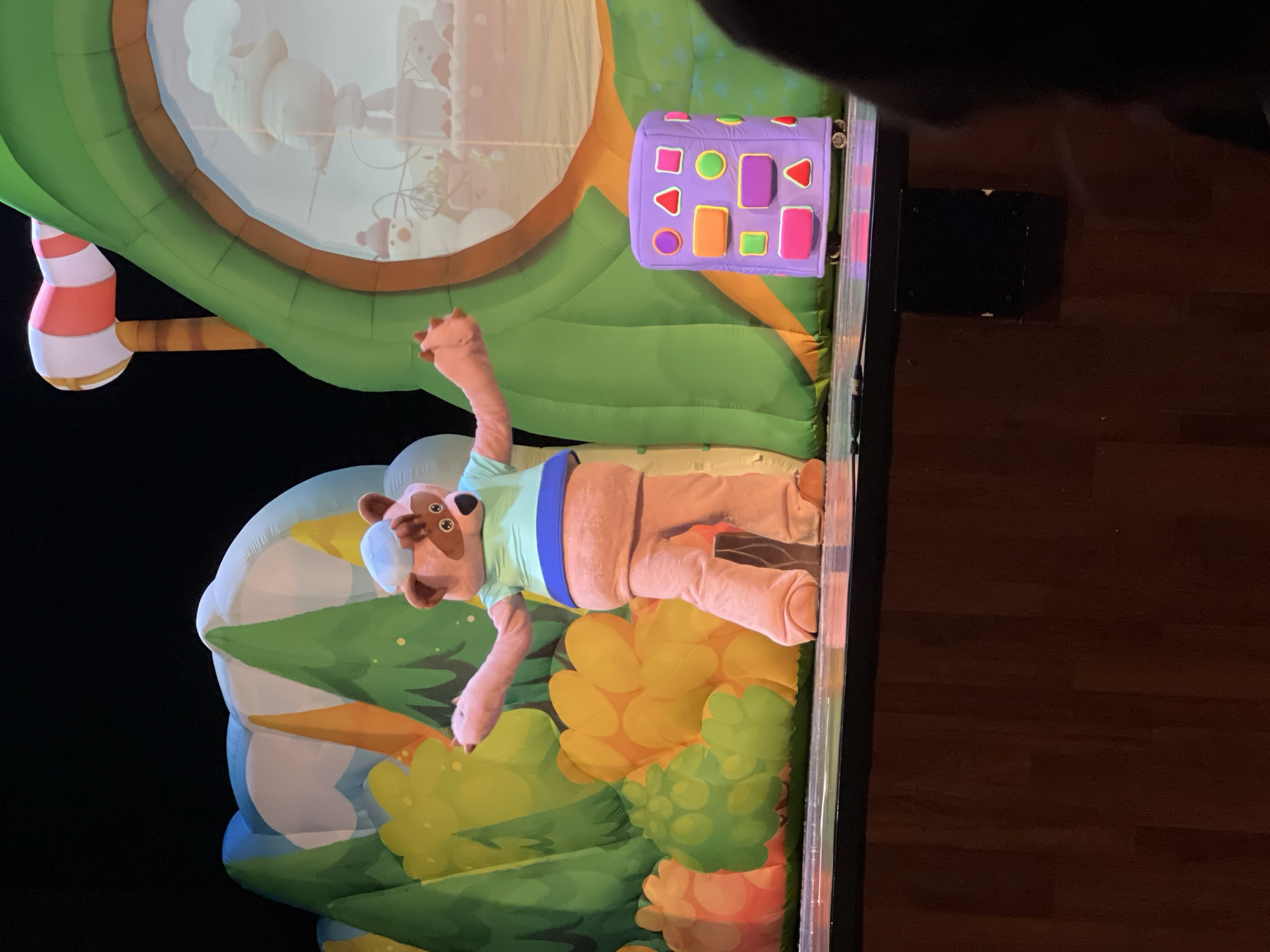 The image depicts an indoor scene with a puppet show set against a black backdrop, possibly in Chaikovsky, Russia (based on the prompt). The show features a pink puppet resembling a bear wearing a light blue shirt with a blue band around its midsection. The puppet is in a lying position with its arms raised. The stage design includes inflatable elements such as a green tree-like structure with a white candy cane stripe, a window displaying cartoon images, a purple toy-shaped block with colorful geometric figures, and a colorful floral background. The scene is illuminated with spotlights. To the right of the set is a dark, likely wooden floor and a black rectangular object on the wall.
