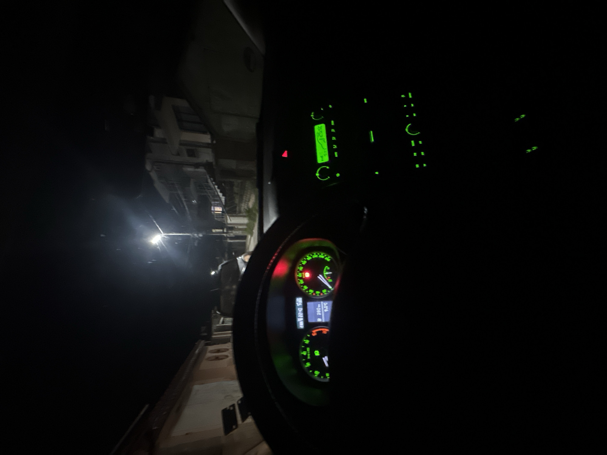 The interior of a vehicle, specifically the driver's area, is visible in the foreground, showing a steering wheel and dashboard illuminated with green lighting. The vehicle appears to be located in what might be an industrial or port area, as evidenced by the exterior view through the window, which shows a structure, potentially a ship or a warehouse, and a light source illuminating the scene. A person is visible outside the vehicle, seemingly standing on a raised platform or walkway. The location is unspecified, described only as the city of Unknown, Unknown.