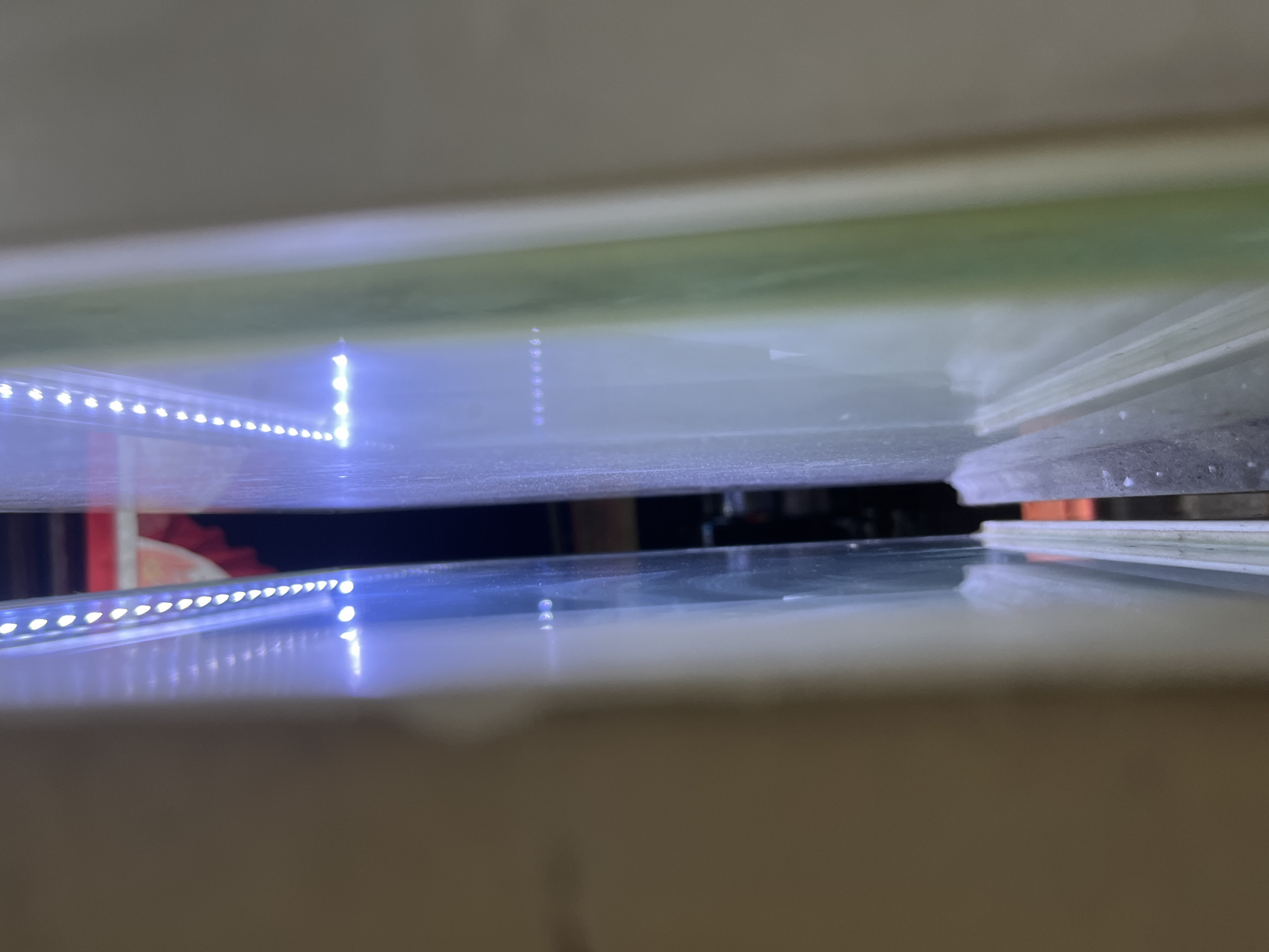 The image shows a close-up, vertical view of two panes of glass, illuminated by a strip of LED lights along the edge of each pane. The glass is clear and appears to be part of a display or enclosure. In the background, a blurred red and yellow pattern is visible, suggesting it might be a fabric or decorative element. The overall lighting is cool-toned due to the blue LEDs. The image was taken in Central Jakarta, Indonesia.