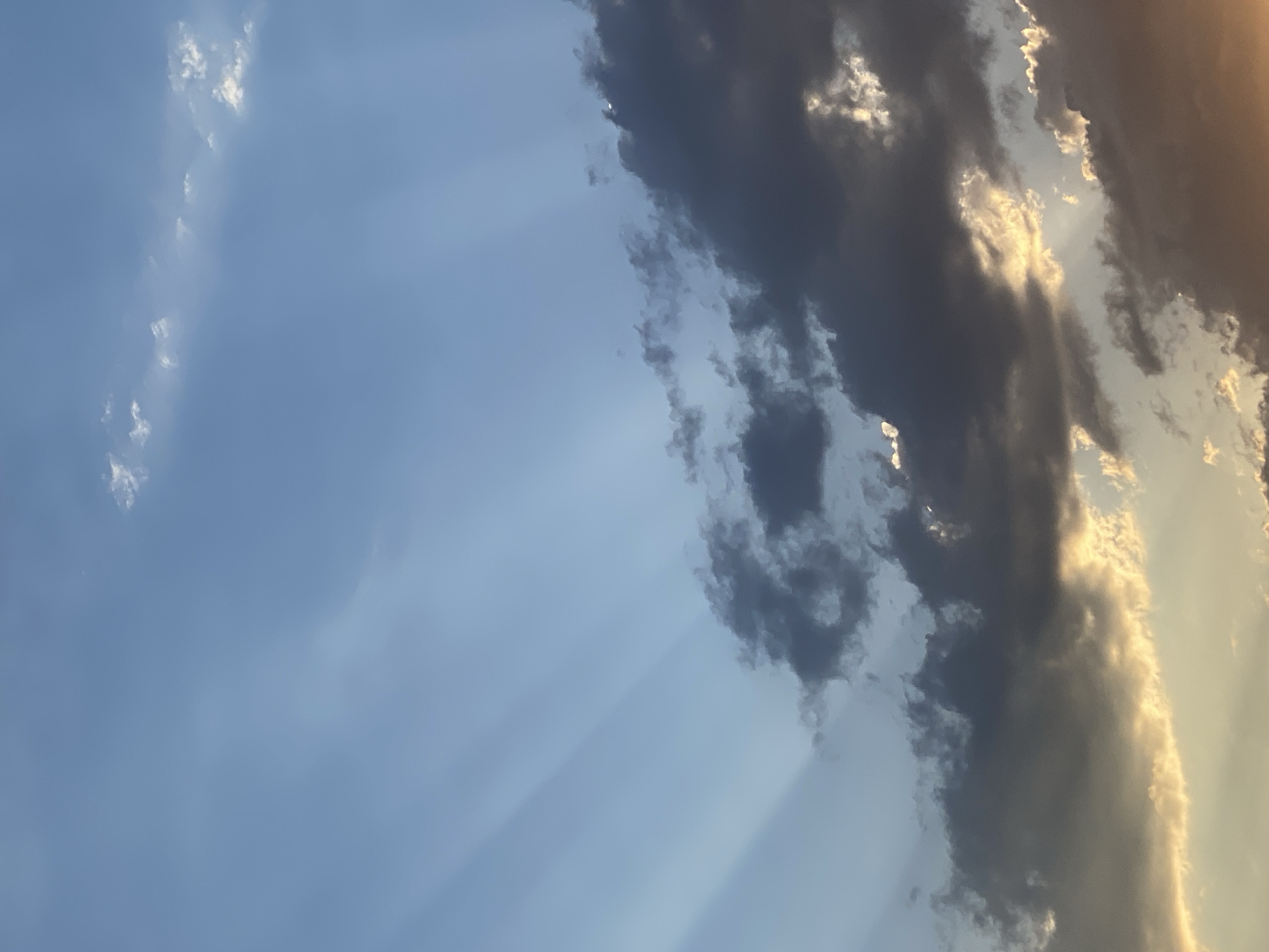 The image shows a sky filled with clouds and crepuscular rays, suggesting a sunset or sunrise. The sky is predominantly blue on the left side, transitioning to darker, heavier clouds on the right, with patches of sunlight breaking through. The crepuscular rays, or sunbeams, are visible as bright shafts of light piercing through the clouds, creating a dramatic effect. The clouds are varied, with some appearing wispy and light, while others are dense and dark, possibly indicating approaching rain or the remnants of a storm. The lighting suggests that the sun is low on the horizon, casting a warm, golden glow on some of the cloud edges. No people, objects, or text are visible in the image. The scene is purely natural, capturing a moment of atmospheric beauty in the sky. Given the location context of Almaty, Kazakhstan, this could be a typical evening or morning sky over the region.