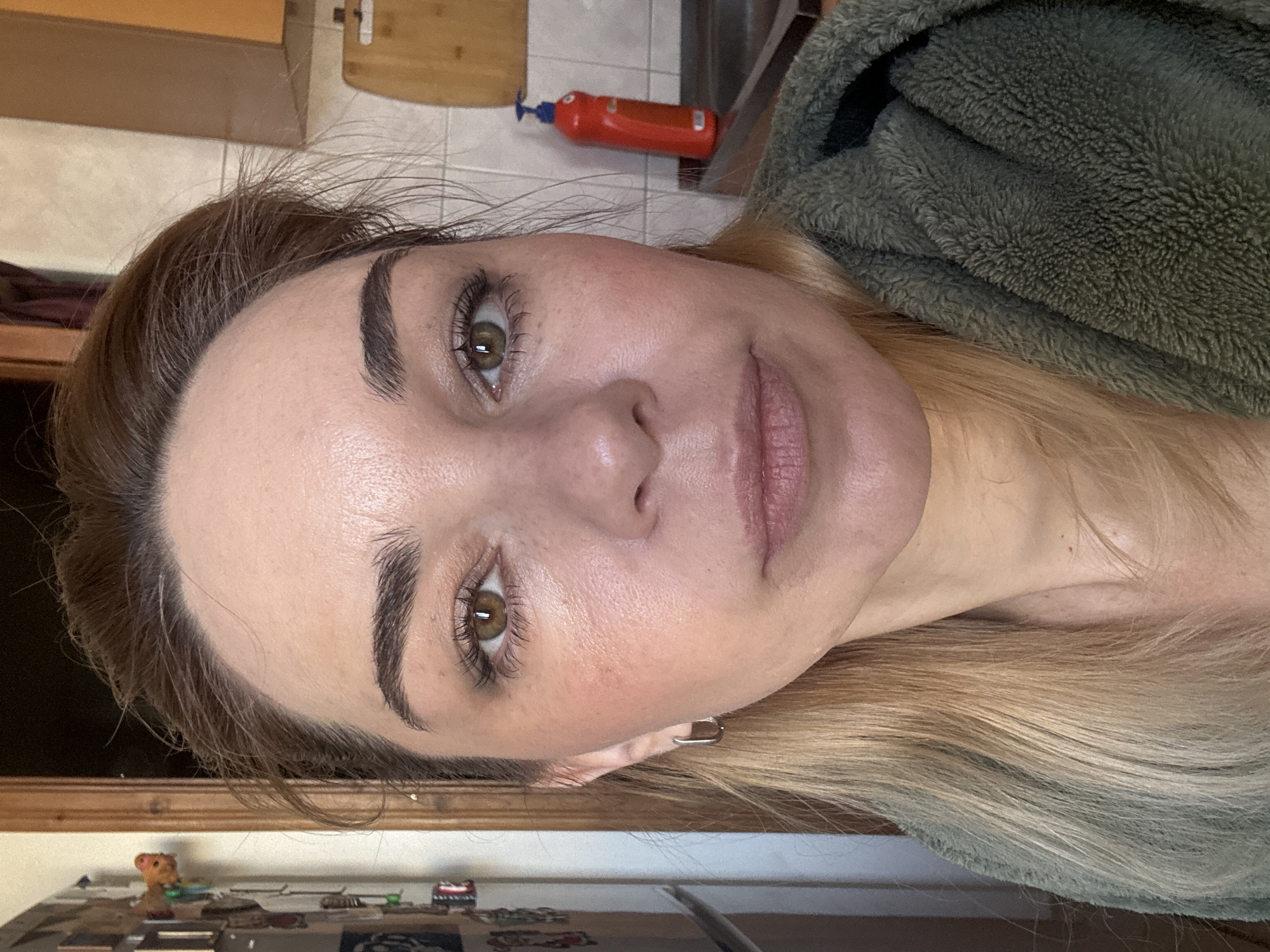 A close-up, angled portrait of a woman with fair skin, brown hair at the roots transitioning to blonde, and hazel eyes. She has well-defined eyebrows and appears to be wearing minimal makeup. The woman is wearing a textured, possibly fleece, green garment.
The background includes elements of an interior, such as wooden cabinets and a tiled wall with a red object possibly a fire extinguisher. The location is unidentified as Unknown, Russia. The photograph does not provide enough information to confirm a specific city or location.
