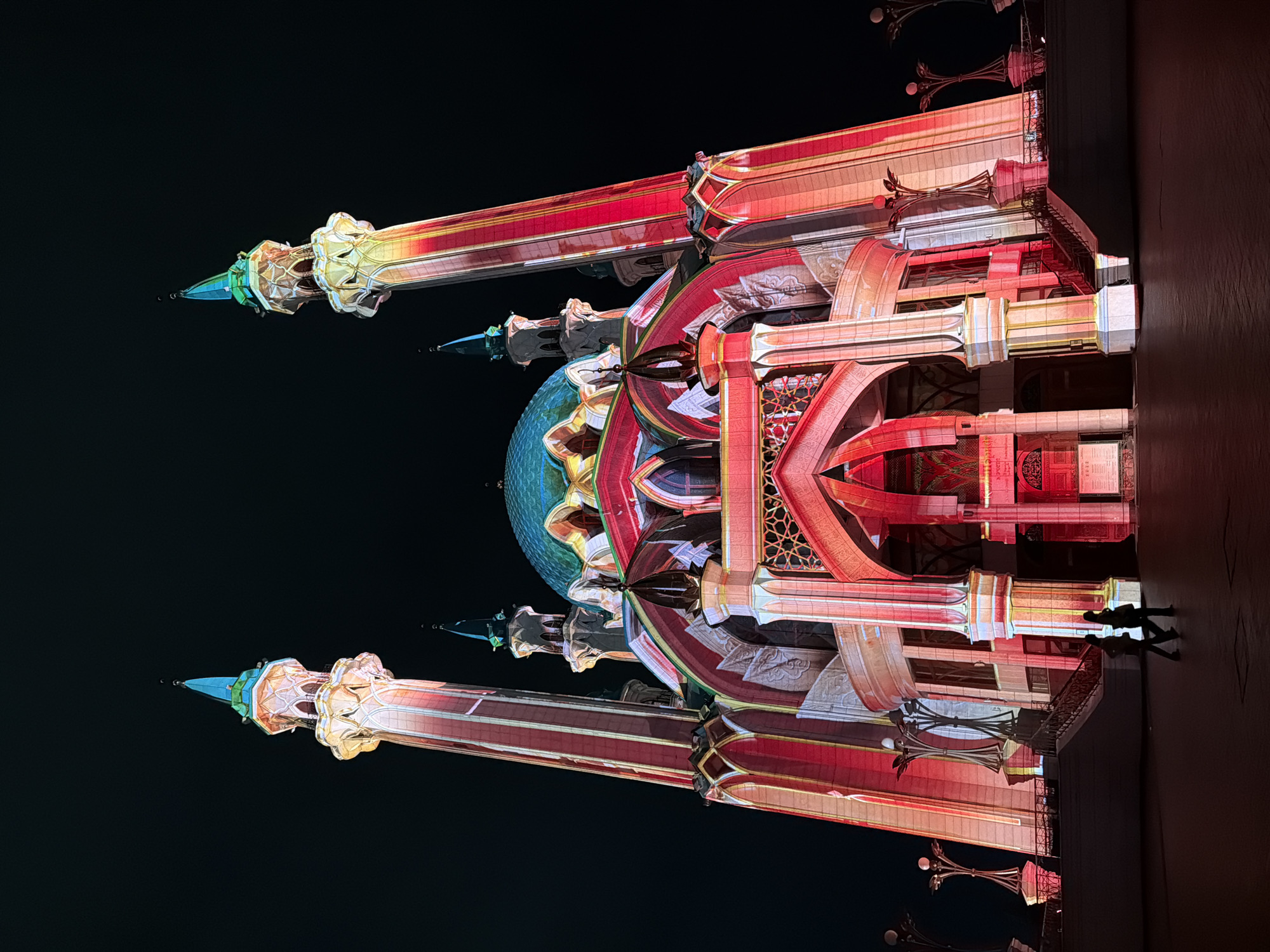 Here is a description of the image:
The image depicts a brightly lit mosque at night. The mosque is illuminated with red, white, and gold colors. It has a blue dome and two minarets. The background is black. There are shadows of people on the lower right. The location is not specified.
