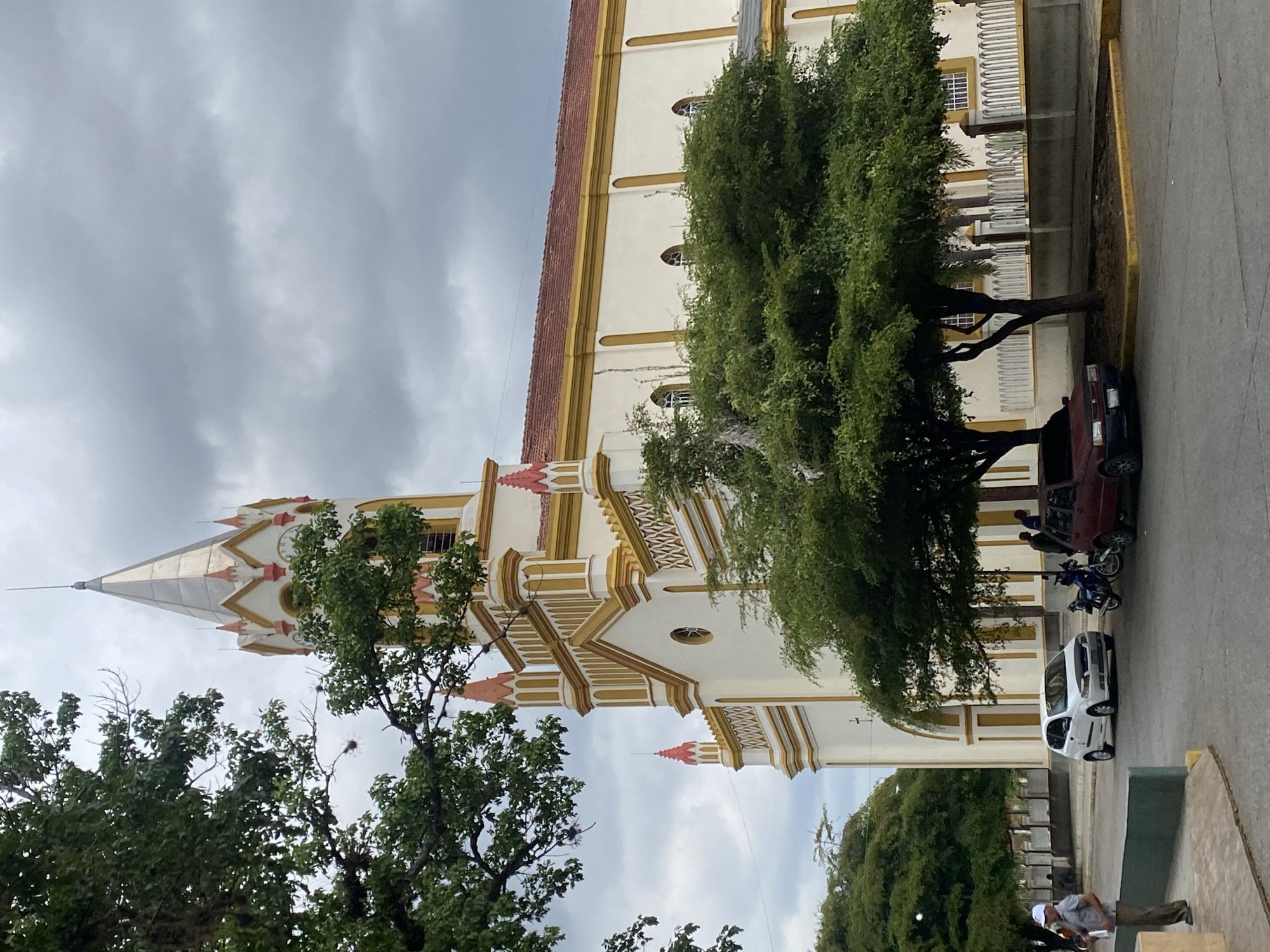 The image shows a tall, ornate church in Tinaquillo, Venezuela, seen from a low angle. The church is white with gold and red trim, and has a spire with a clock. Trees partially obscure the church's facade. A street runs along the side of the church with a maroon car and a white car parked on it, as well as a motorcycle and two people standing near the maroon car. There are dark clouds in the sky. A person is sitting to the right in the bottom corner.