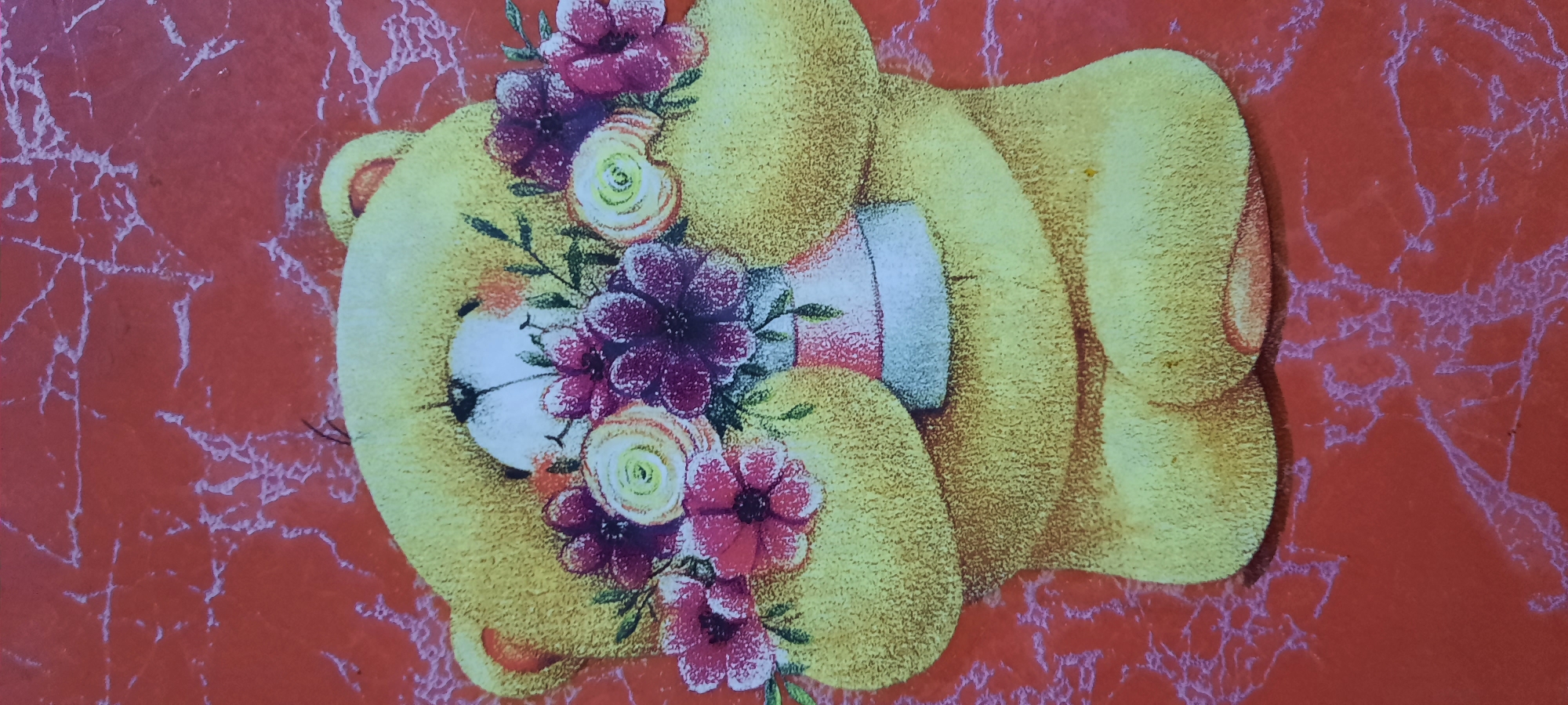 A close-up, top-down shot shows a yellow teddy bear doll holding a bouquet of flowers against a red, textured background. The teddy bear's head is tilted down, obscuring its face behind the flowers, which include pink, purple, and yellow blossoms with green leaves. The red background features a lighter, vein-like pattern. The image appears to be a decorative item, possibly a sticker or a printed card, and is not set in any specific urban location.