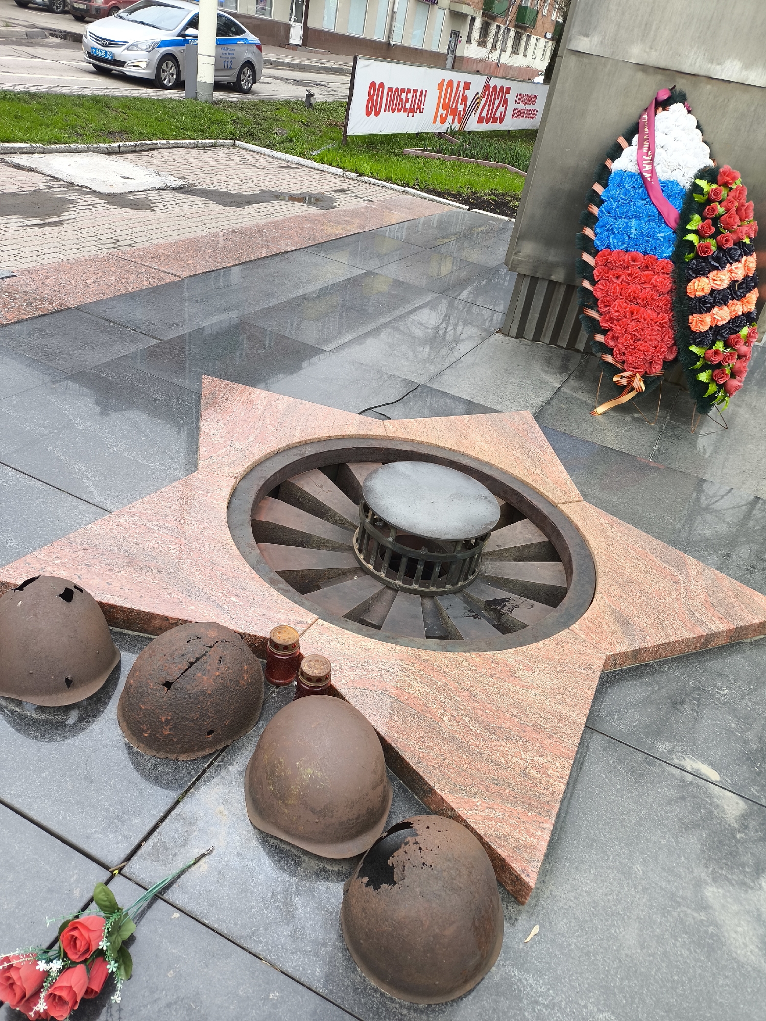 The image depicts a war memorial in Khimki, Russia, specifically a monument commemorating the Great Patriotic War. The memorial features a five-pointed star made of red granite, housing an eternal flame. Around the base of the star are arranged several heavily damaged military helmets, likely Soviet-era. A small bouquet of red flowers and several candle holders also lie near the helmets.
Behind the star, attached to a gray concrete structure, is a large wreath decorated with the colors of the Russian flag: white, blue, and red. A banner in the background displays the text "80 Victory! 1945-2025". A police car is visible parked nearby, indicating the location is in a public space. The ground is paved with gray tiles, some areas appearing wet.