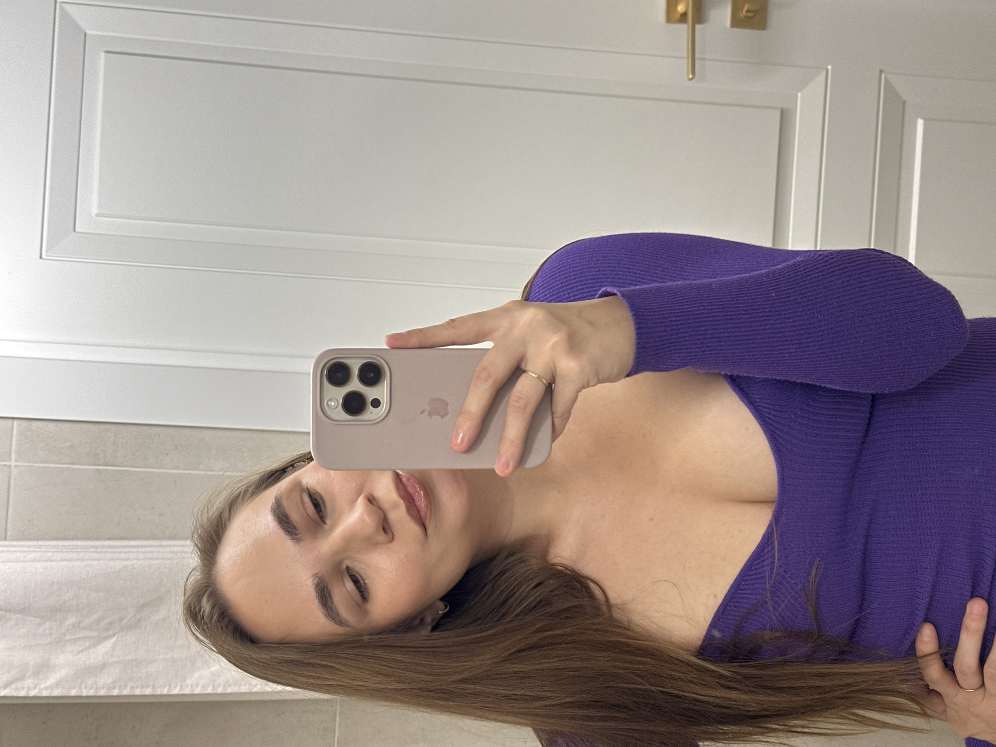 A woman with brown hair is taking a selfie in a bathroom. She is wearing a purple, low-cut sweater. She is holding a light purple iPhone in her right hand to take the photo. Behind her is a white door with gold hardware, and a grey tiled wall and a white towel are visible in the lower left. The location, Kharnavy, Russia, cannot be verified from the image.