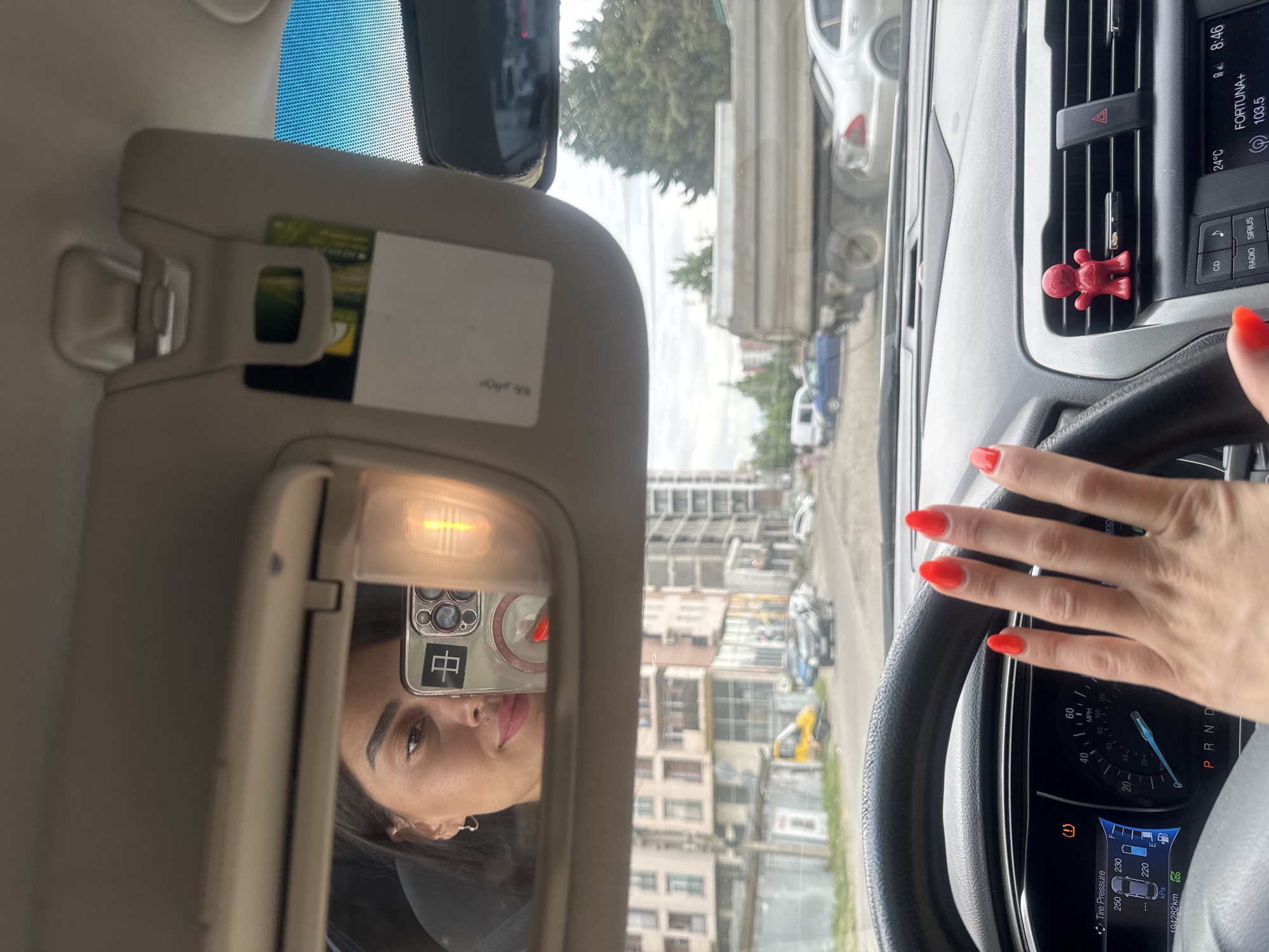The image shows a selfie taken by a woman inside a car, likely in Tbilisi, Georgia. She is visible in the car's vanity mirror, looking at the camera while holding up her phone to take the picture. The woman has dark hair, dark eyebrows, and is wearing makeup, including bright red lipstick and matching red nail polish. Her left hand, also with red nail polish, is extended towards the steering wheel.
The car's interior is visible, including the steering wheel, dashboard with a speedometer and other gauges, and a vent with a small red novelty air freshener clipped to it. The dashboard also shows a "P" for park on the gear selector.
Outside the car, through the windshield, a street scene in a city is depicted. There are multi-story buildings, a yellow truck, a silver dump truck, and several cars. The presence of these vehicles and buildings suggests an urban environment. The overall lighting indicates it is daytime. The image does not provide specific landmarks to confirm it is Tbilisi, Georgia, but it is consistent with an urban setting in that region.