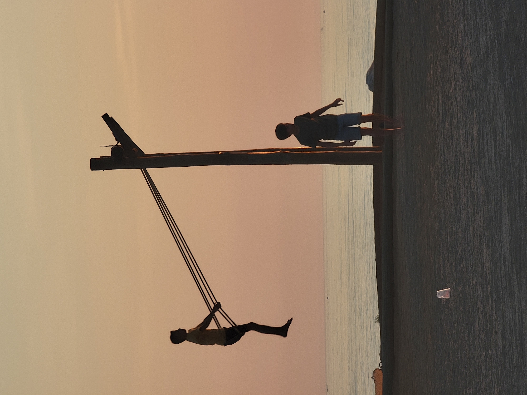 At sunset on Quarteira beach in Portugal, a boy swings high on a rope swing attached to a wooden pole structure. His silhouette is stark against the pale orange sky as he hangs suspended in the air, his body arched. To his right, near the base of the pole, another boy stands looking out towards the ocean. The water is calm, reflecting the soft hues of the sky. The ground is sandy, with a discarded plastic cup visible in the foreground. The scene captures a moment of youthful joy and freedom by the sea.