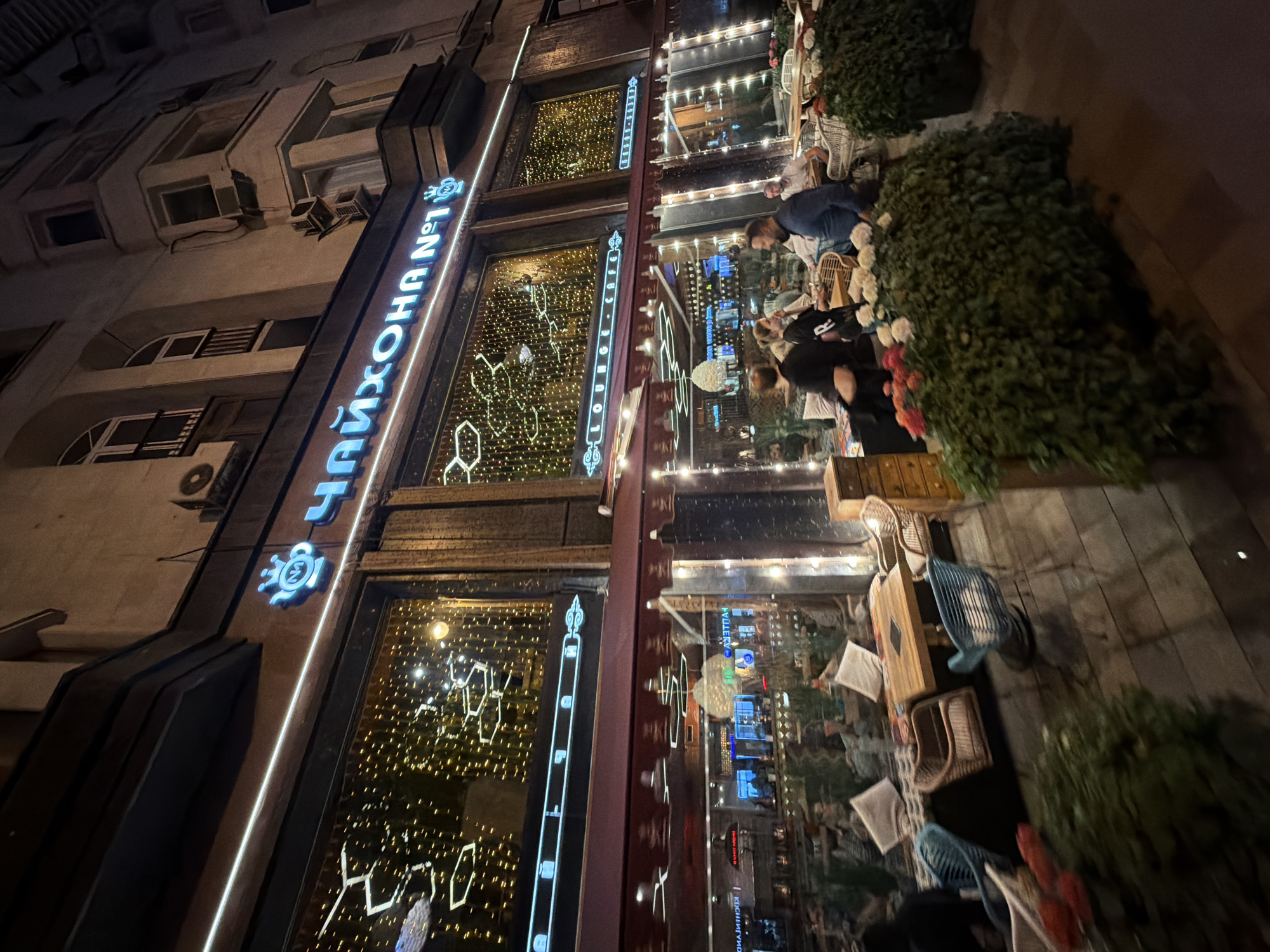 The image shows the exterior of "Chaihona No. 1 Lounge Cafe" in Moscow, Russia, at night. The cafe's name is prominently displayed in illuminated Cyrillic letters. The facade features large windows decorated with patterned lights. Outside, there is outdoor seating with tables, chairs, and greenery. People are sitting and interacting at the tables. The building is multi-storied and has residential elements.