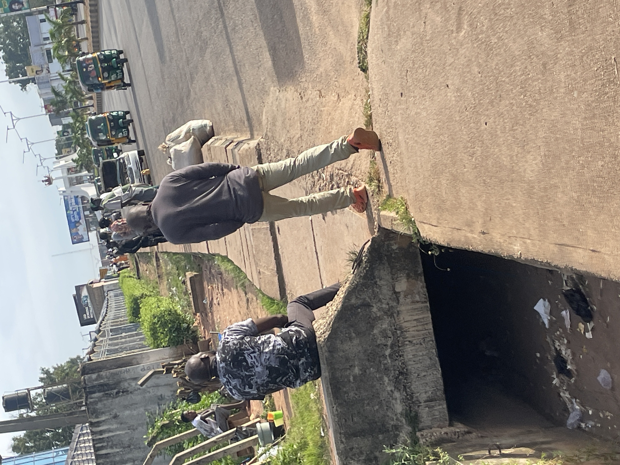A medium-shot, eye-level, outdoor photograph taken on a sunny day shows a street scene in Jos, Nigeria. In the foreground, a man is seated on a concrete structure, his back to the camera. He is wearing a dark, camouflage-patterned t-shirt and dark trousers. To his right and further down the concrete pathway, another man is standing, facing away from the camera, wearing a grey long-sleeved shirt and light-colored trousers. He has a large, white bag on his shoulder. In the background, there is a street with several auto-rickshaws, also known as Keke Napep, in motion. The environment appears to be urban, with buildings, trees, and streetlights visible along the roadside. There is also a drainage ditch in the lower left portion of the frame, containing debris.