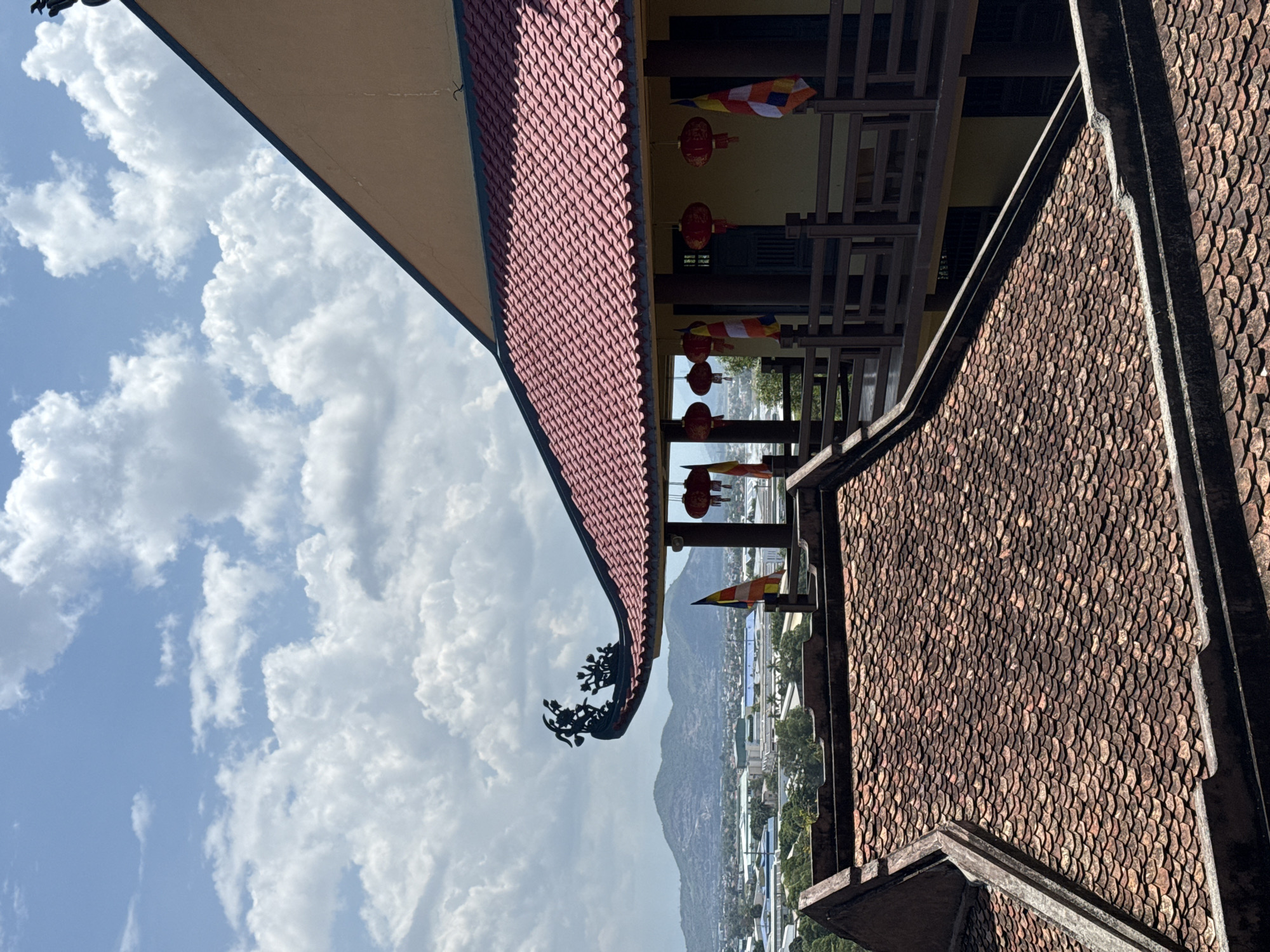 The photograph shows a view of Tay Nha Trang Ward, Vietnam, through the architectural elements of a Buddhist temple. In the foreground, sections of the temple roof are visible, featuring intricate red and brown tiling. A decorative black ornament adorns the edge of the roof, contrasting with the sky. Hanging red lanterns and multicolored flags are suspended, adding color to the structural details of the temple. Beyond the temple, a mountain range rises against a sky filled with puffy white clouds. The buildings of Tay Nha Trang Ward stretch across the cityscape in the distance.