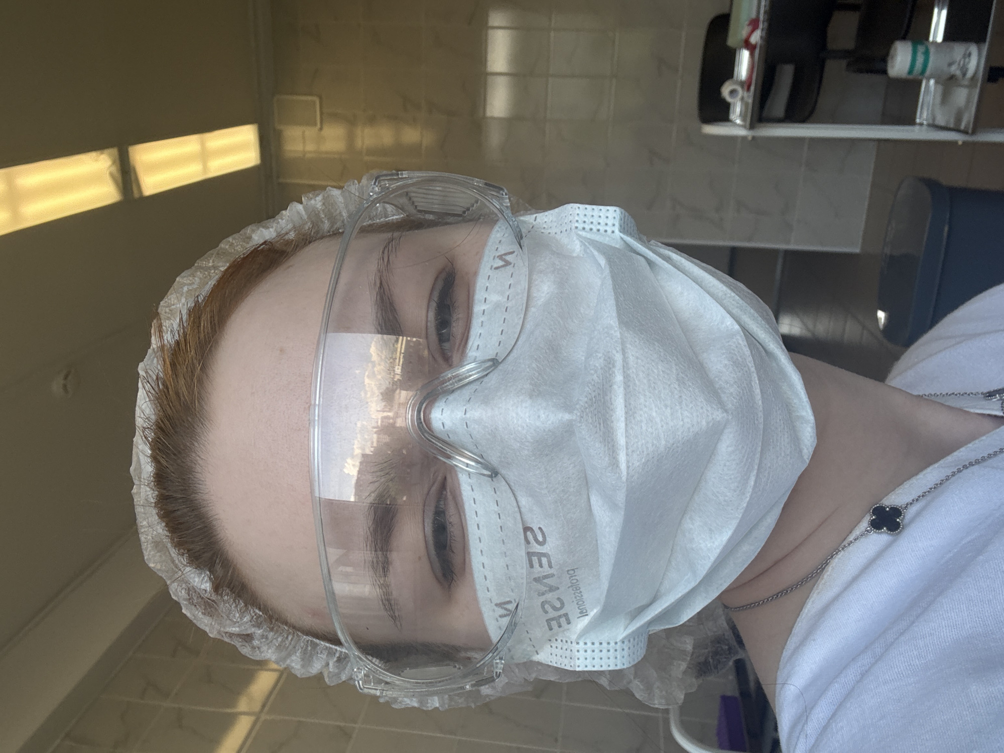 A self-portrait captured in Saint Petersburg, Russia, shows an individual wearing personal protective equipment (PPE) in what appears to be a medical or laboratory setting. The subject, seen from the chest up, has reddish-brown hair mostly covered by a disposable white bouffant cap. They are wearing clear plastic safety glasses, which reflect bright horizontal fluorescent lights from the upper left. A white disposable face mask covers the lower half of their face.
The person's eyes, with dark eyelashes and eyebrows, are visible above the mask. Their gaze is directed straight towards the camera. The mask has visible reversed text, clearly reading "SENSE" and "Professional" in dark print. The individual is wearing a white collared shirt, and a silver chain necklace with a black four-leaf clover (quatrefoil) pendant hangs around their neck.
In the background, light-colored tiled walls are visible, along with reflections of what seem to be ceiling lights. To the right, a shelf holds various items, including what looks like rolls of paper or medical supplies, suggesting an organized, functional environment. The overall impression is one of professionalism and adherence to safety protocols.