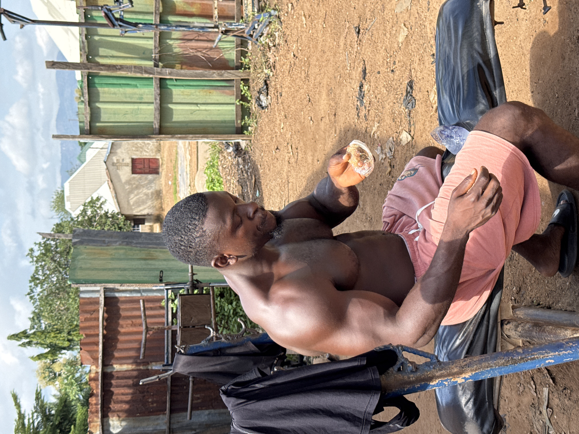 The image depicts a muscular Black man, shirtless and wearing pink shorts, sitting on a blue metal structure outdoors. He is holding a piece of food in his right hand, bringing it towards his mouth. A black garment, possibly clothing, is draped over the structure in front of him. The background shows a rough, earthy ground with scattered debris, suggesting an unpaved area. Behind him, there are various structures, including corrugated metal sheeting in green and rusted brown, and a small building with a red door. Trees and vegetation are also visible, along with a cloudy sky, hinting at daylight. The overall setting appears to be a somewhat rudimentary outdoor space, possibly a yard or a training area, in Akwanga, Nigeria. The man's physique suggests he might be involved in physical activity or training.