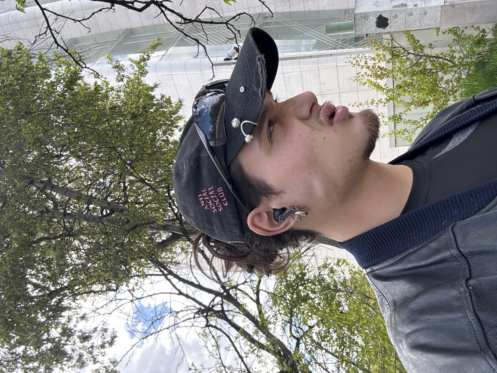 The image displays a close-up, vertical portrait of a young man, rotated 90 degrees clockwise, looking upwards. He is positioned outdoors against a backdrop of trees, a modern building, and the sky.
The man has a light complexion, dark hair, and a short beard outlining his jawline. He is wearing a black baseball cap with the brand "ANTI SOCIAL SOCIAL CLUB" printed upside down in pink lettering on the front. A pair of black sunglasses, with the word "Bolle" visible on the arm, are tucked into the cap's brim. The cap also features several metallic adornments, including a screw and a silver, double-hoop piercing-style ornament on the side. A black earbud is visible in his right ear, along with a silver hoop earring in his earlobe. He wears a black t-shirt beneath a dark blue or black jacket that appears to be leather or a similar material, featuring a ribbed collar. His mouth is slightly open, and his gaze is directed upwards, suggesting observation or contemplation.
The background features vibrant green trees with delicate branches, indicative of spring or summer foliage. Beyond the trees, a modern, light-colored building with a sleek, multi-layered design and reflective surfaces is partially visible. The sky is bright with some clouds. The natural light suggests it is daytime. The overall scene indicates an urban environment with green spaces, likely in Sofia, Bulgaria.